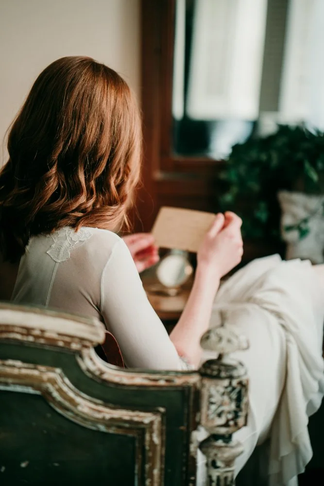 A woman with wavy reddish-brown hair sitting on an antique carved wooden chair, reading a letter near a window with sunlight and a houseplant in the background.