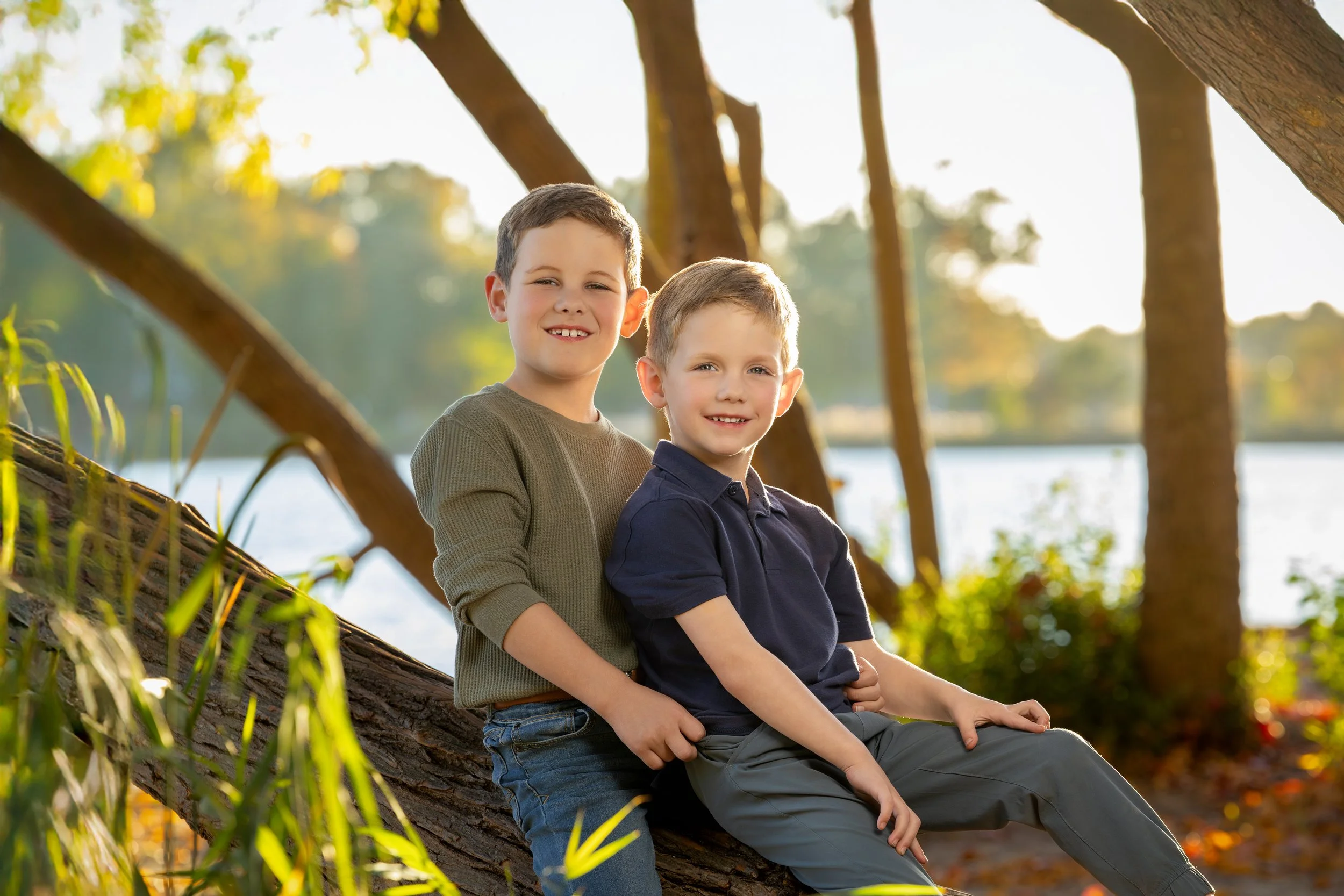 Two young boys sitting on a fallen tree by a body of water, smiling at the camera, with trees and sunlight in the background.