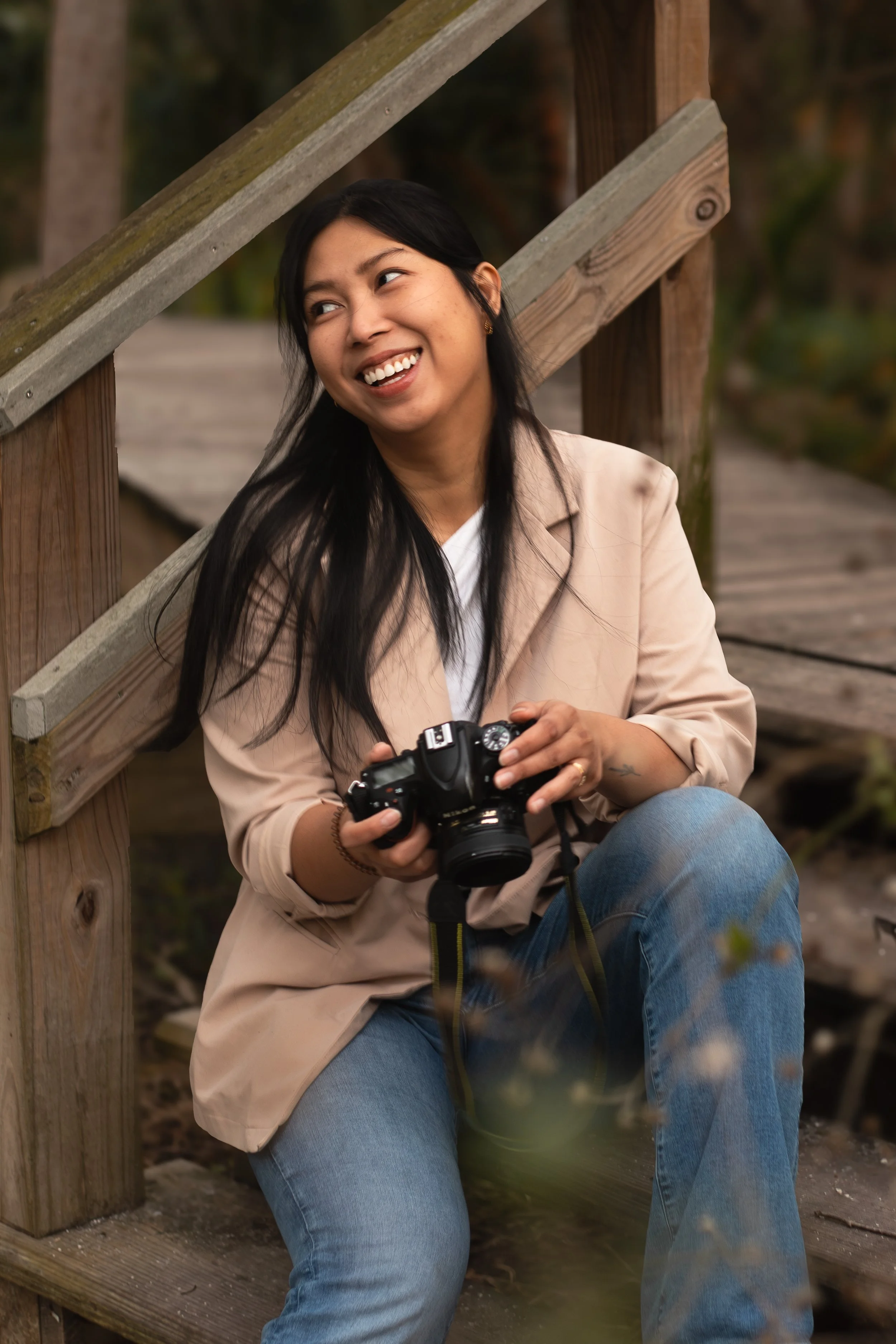 A woman sitting on a wooden bridge, smiling and holding a camera.