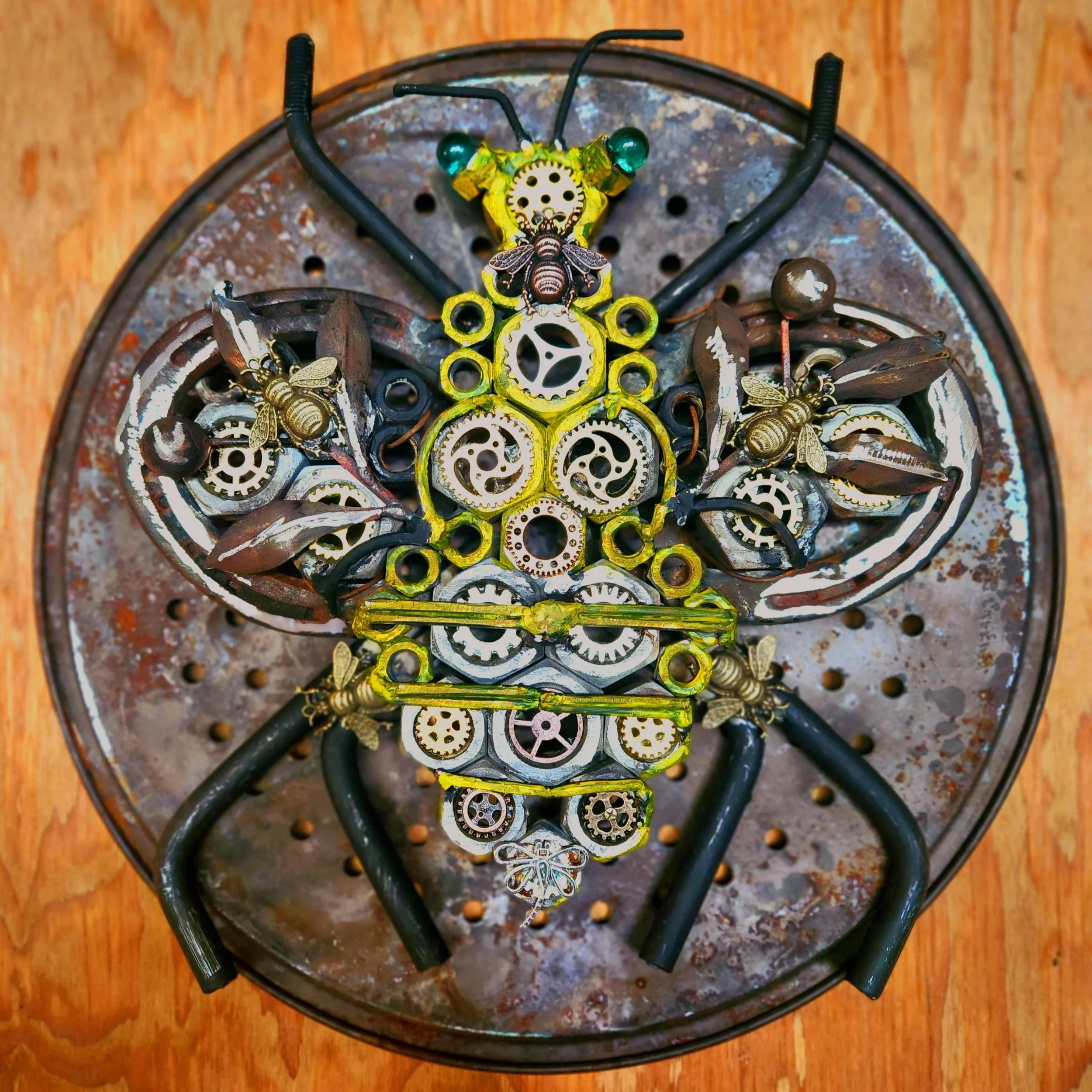 A bee sculpture made of small gears, metal parts, and insect-shaped objects, assembled to resemble a flying insect, placed on a round perforated metal surface.