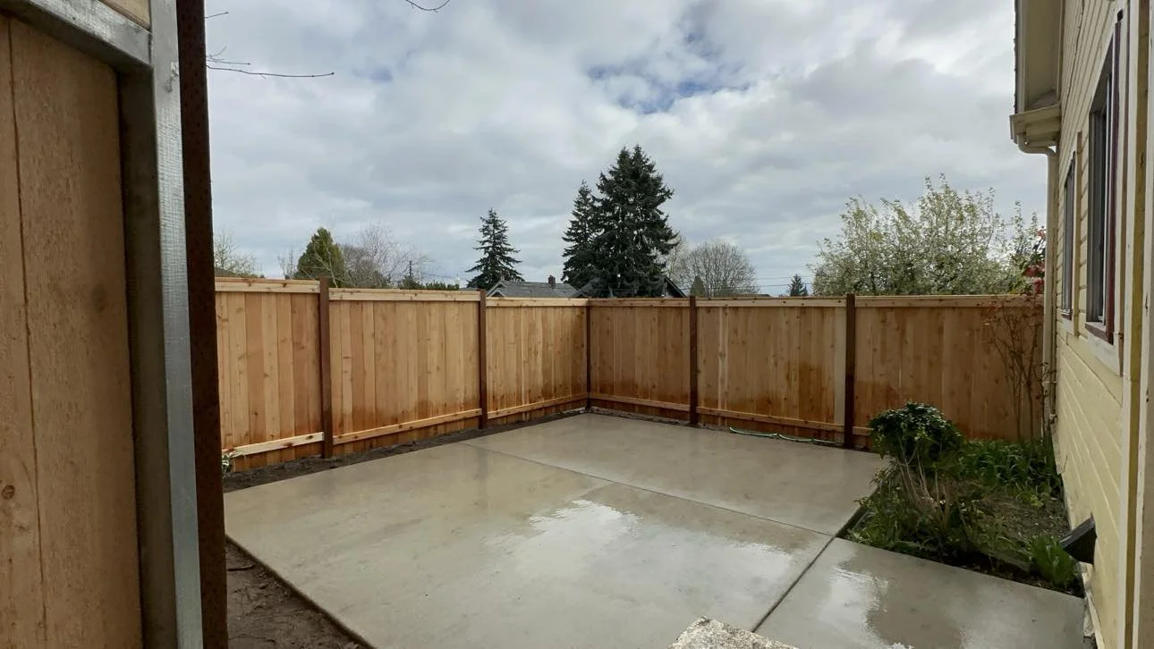 A backyard with a freshly poured concrete patio surrounded by a wooden fence, with a house on the right and some small plants in a garden bed in the corner.