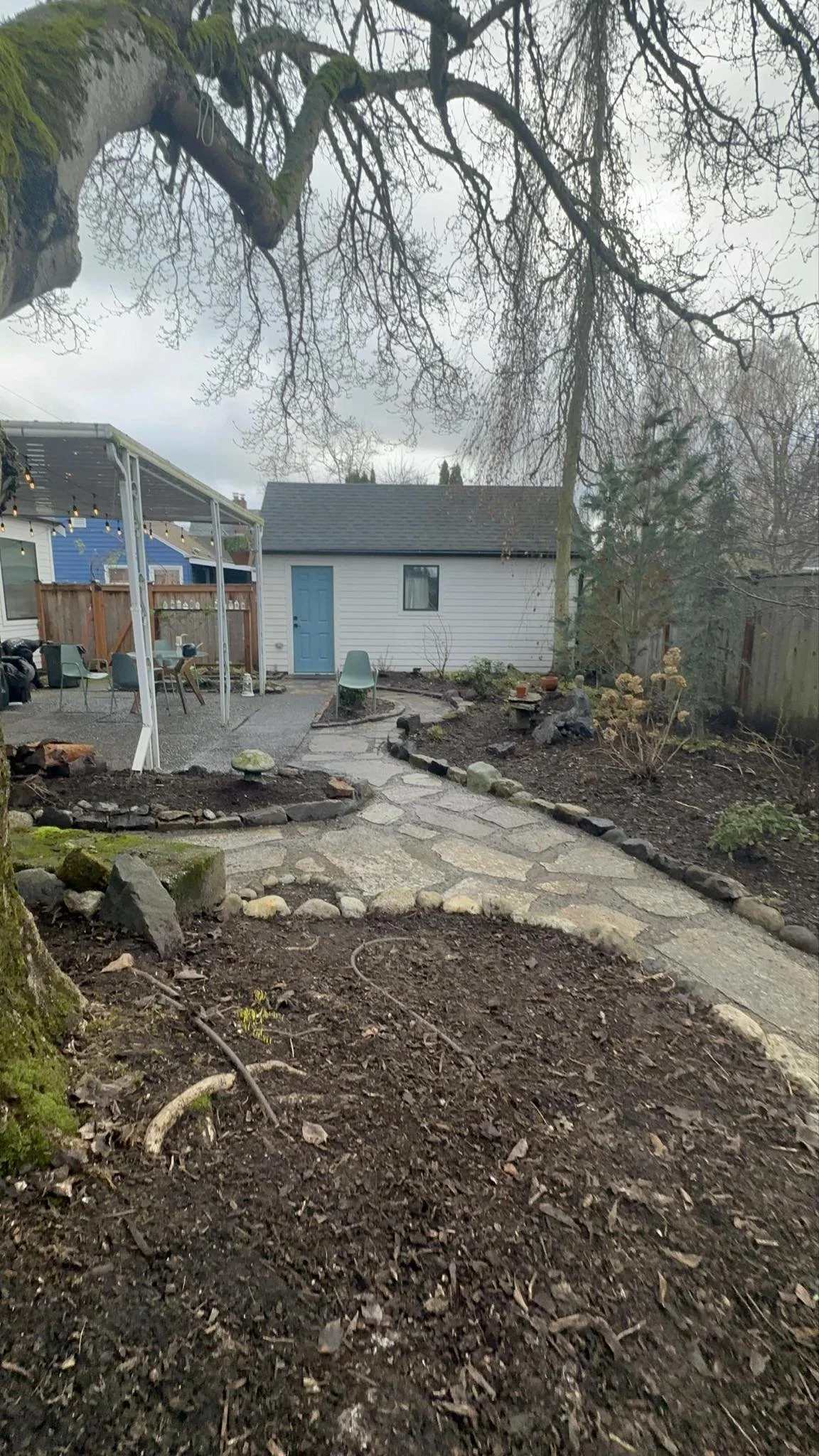 Backyard with a winding stone pathway, trees, a blue and white shed, patio area with seating, and string lights