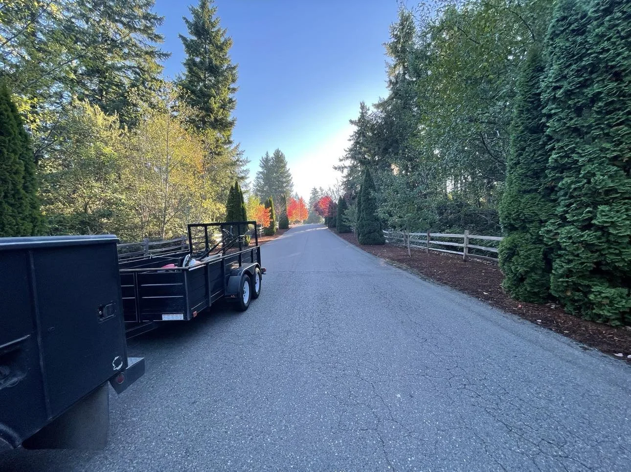 Empty paved rural road lined with green trees and shrubs, with a black trailer parked on the left side.