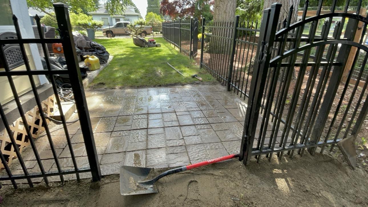 Outdoor scene showing a partially fenced backyard with a new stone patio, grass, a lawn mower, a yellow gas can, and gardening tools, with a house and trees in the background.