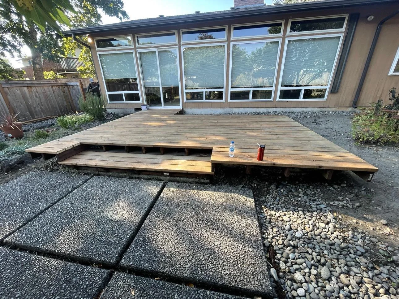 Newly built wooden deck attached to a house, with construction tools and a water bottle on it, surrounded by rocks and a paved patio, in a backyard with plants and a wooden fence.
