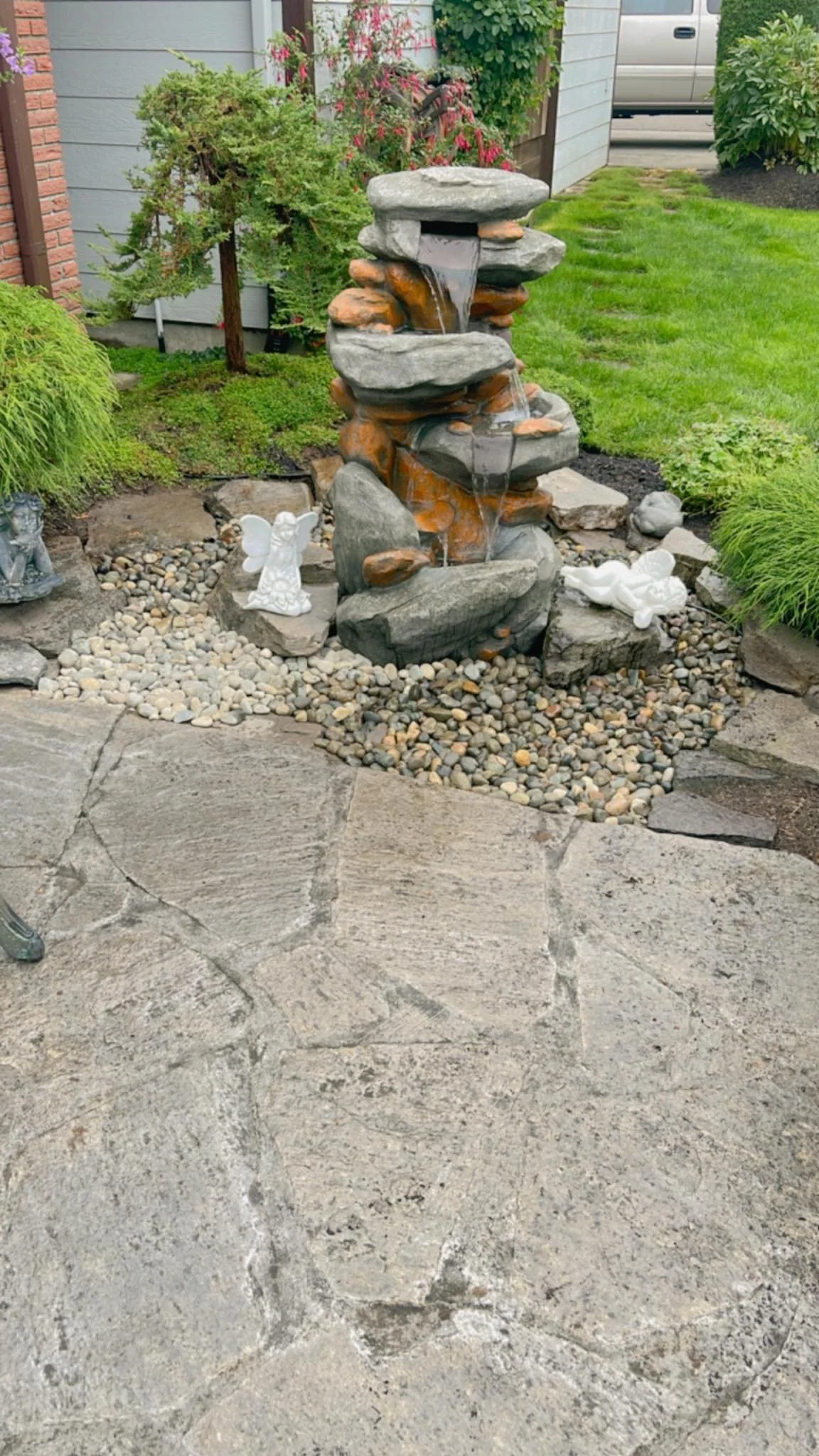 A small outdoor garden fountain with water flowing over stacked rocks, surrounded by decorative angel statues, pebbles, and lush greenery on a stone patio.