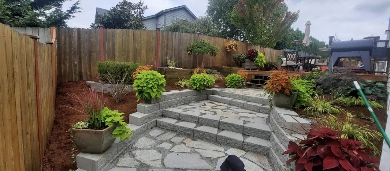 A backyard garden with stone steps leading up to a patio. The area is bordered by a wooden fence and decorated with potted plants and flowers.