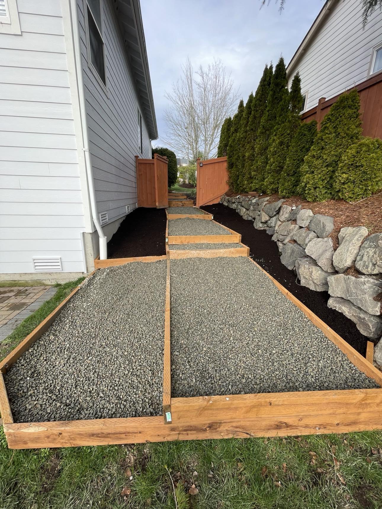 A newly constructed pathway with wooden borders filled with gravel leading away from a house, bordered by a black mulch bed and green shrubs, with a wooden gate at the end.