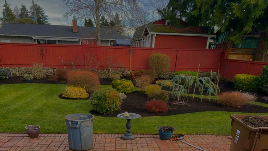 Backyard garden with a brick patio, various shrubs, and trees, enclosed by a red wooden fence.