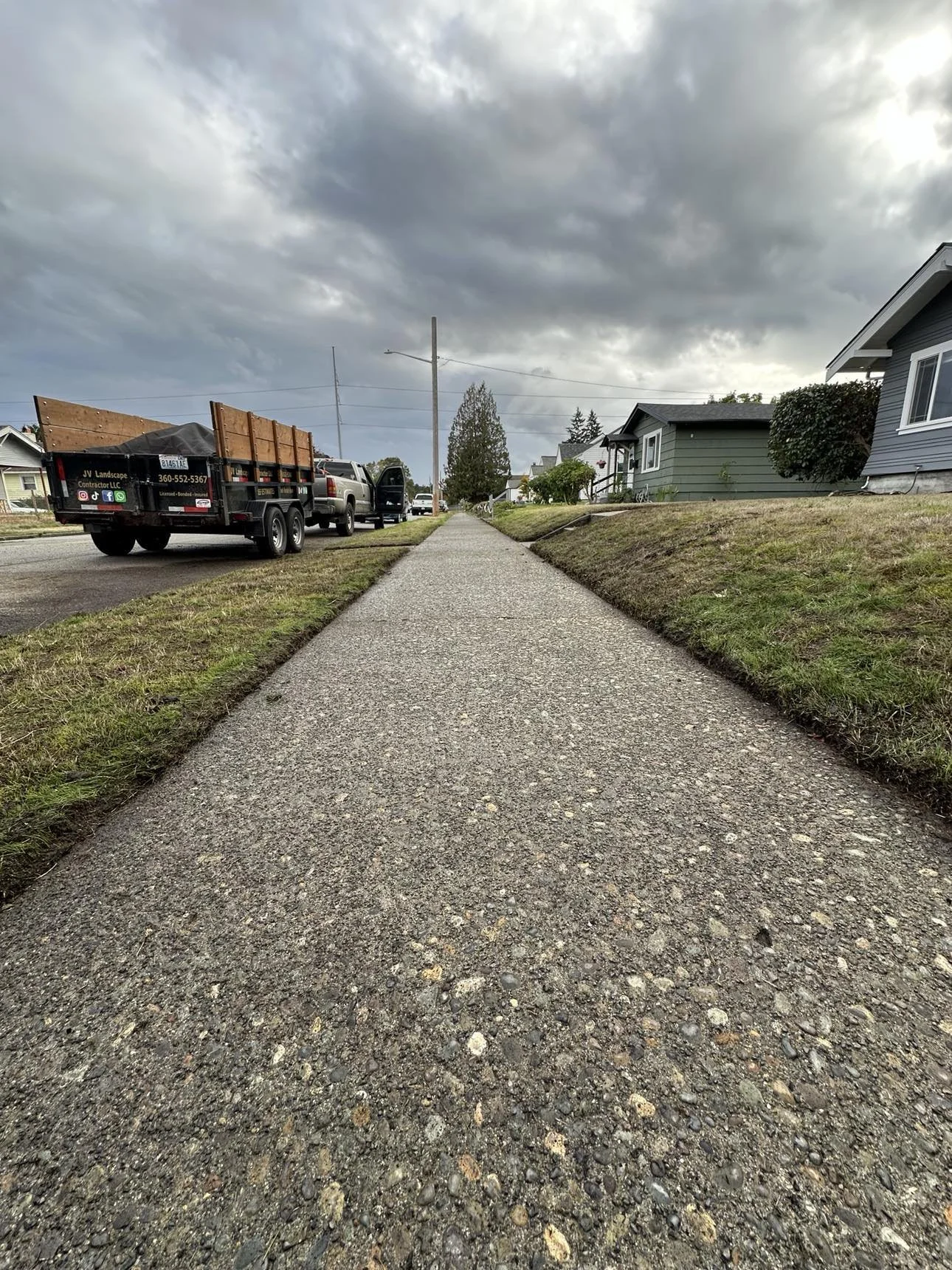 A sidewalk running through a neighborhood with houses on one side and a street with a truck and cars on the other, under a cloudy sky.