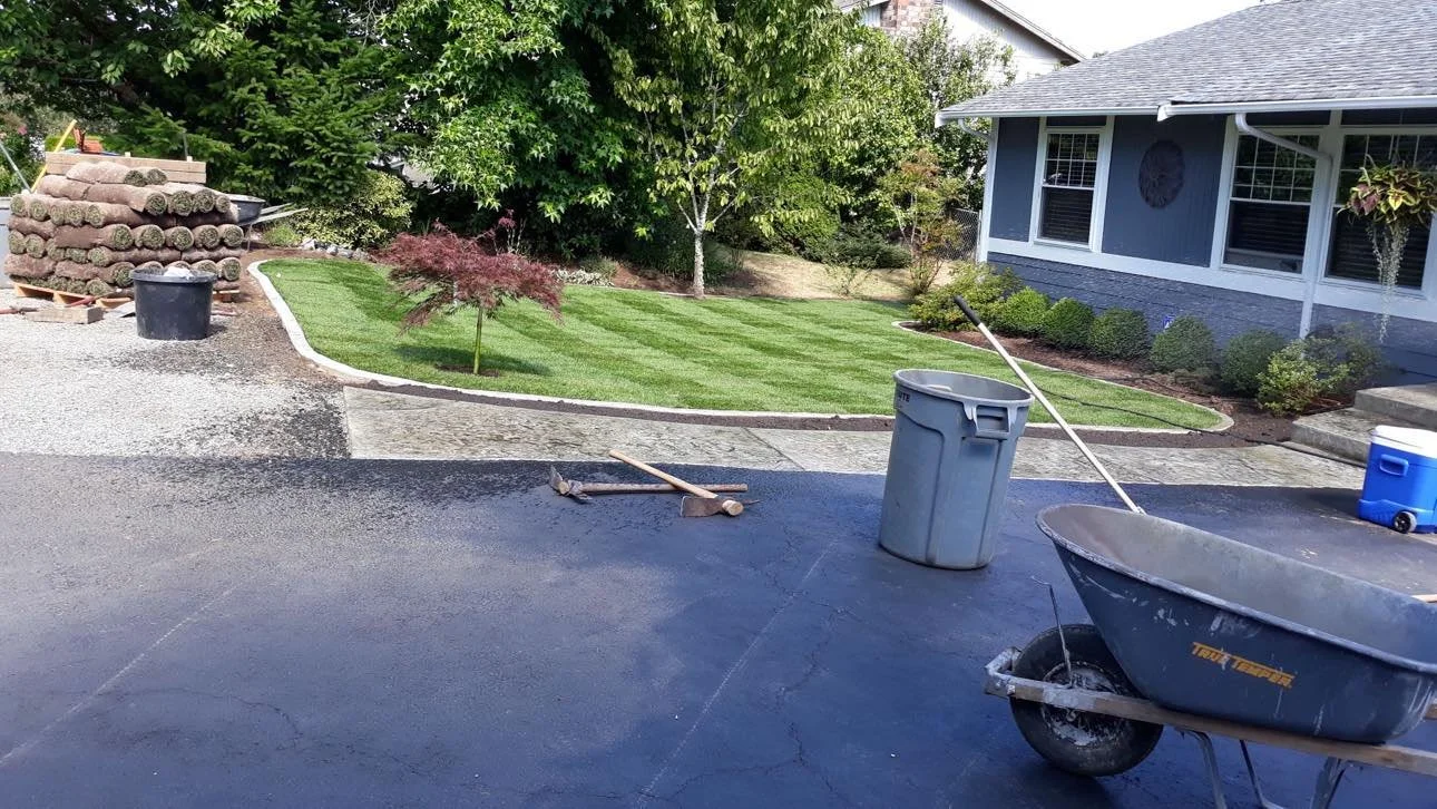 A driveway being resurfaced with asphalt, construction tools, a trash can, a wheelbarrow, and a landscaped front yard with bushes and a tree, next to a house with blue siding and windows.
