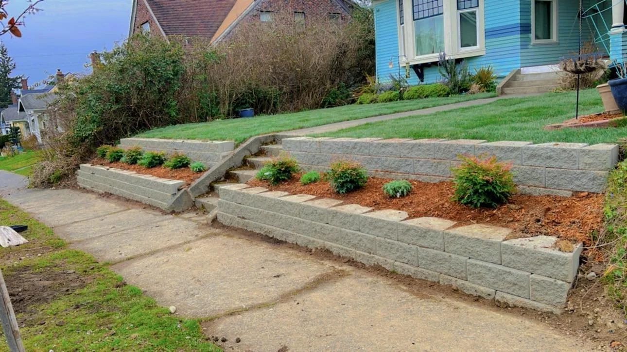 A residential front yard with newly constructed tiered stone retaining walls with staircases, small green shrubs planted in mulched beds, a pathway, and a blue house in the background.