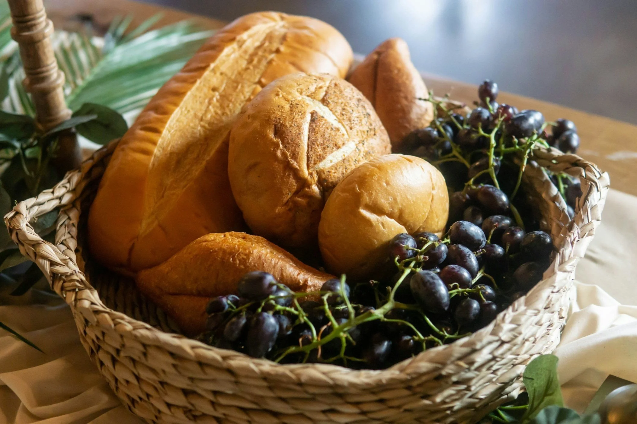 A woven basket filled with various types of bread, including baguette, rolls, and croissants, next to a bunch of black grapes on a table.