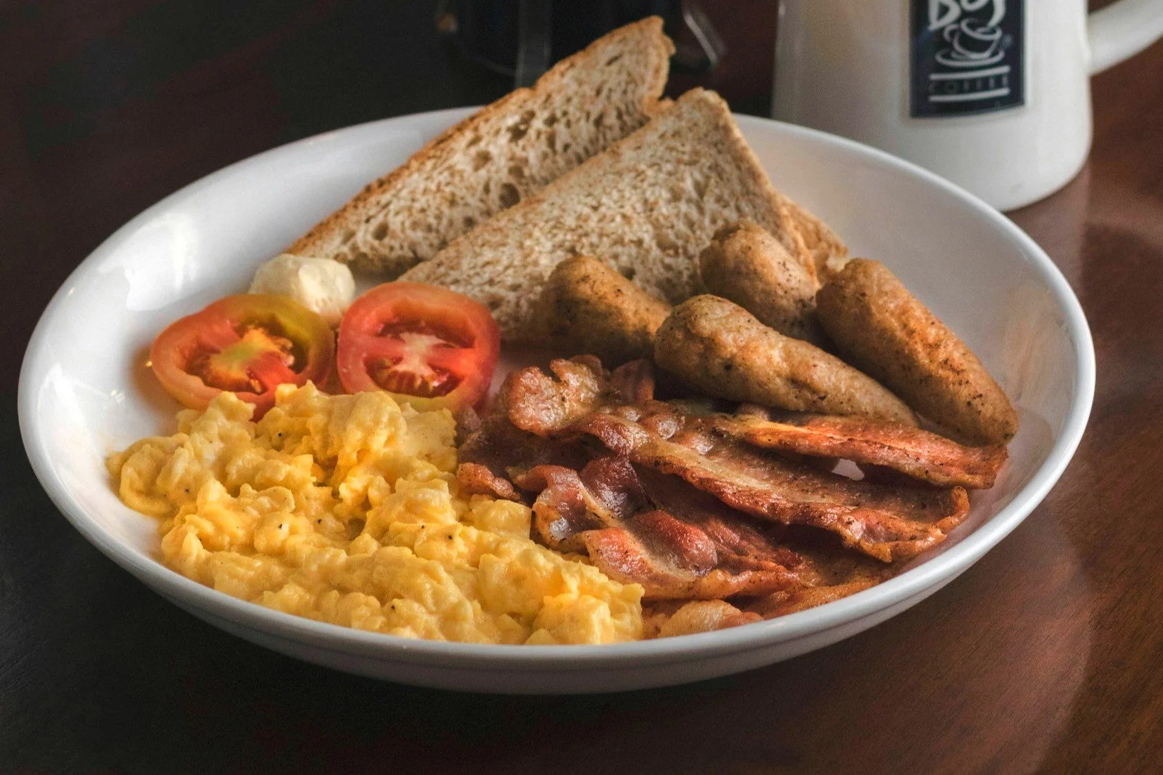 A breakfast plate with scrambled eggs, sliced tomatoes, two slices of whole wheat toast, sausage links, and bacon on a white dish with a coffee mug in the background.