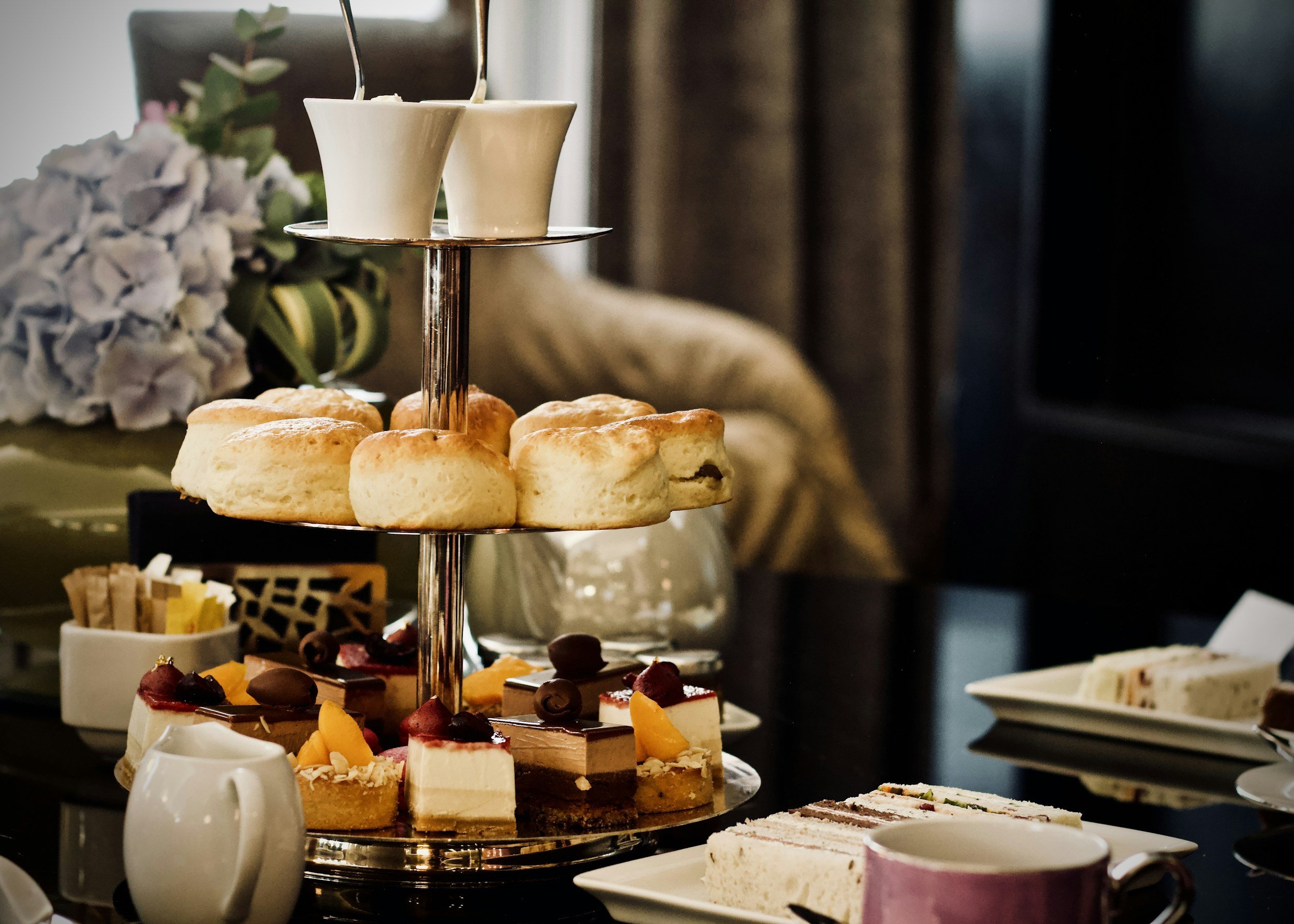 A three-tiered serving tray with assorted scones, mini cakes, and desserts, with plates of sandwiches, cups, sugar packets, and a vase with flowers in the background.