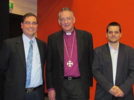 Three men standing together, with the man in the middle wearing a clerical collar and a large cross necklace. The background is red and orange.
