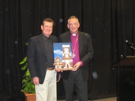 Two men standing on stage, holding a framed picture of a gingerbread house with a tower, in front of a black curtain.