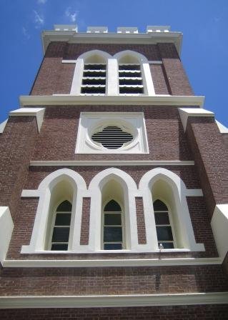 Close-up of a brick church tower with white accents, arched windows, and a louvered vent near the bottom, set against a clear blue sky.