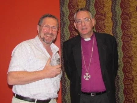 Two men stand together, smiling, with one holding a glass of beer. One is dressed casually, the other in religious attire with a purple shirt and a cross necklace. They are standing indoors in front of a red and patterned curtain.