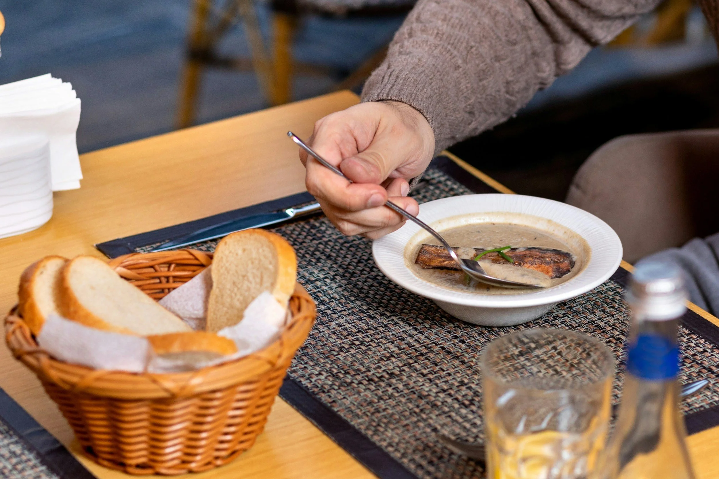 A person holding a spoon and eating soup with pieces of bread in a basket nearby on a dining table.