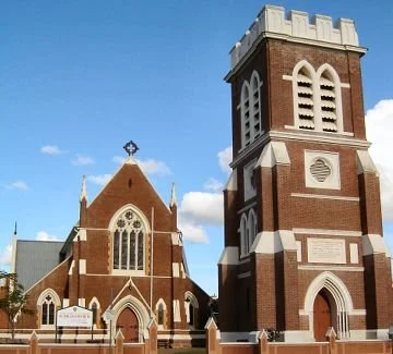 A brick church with a tall bell tower, pointed arch windows, and a blue sky background.