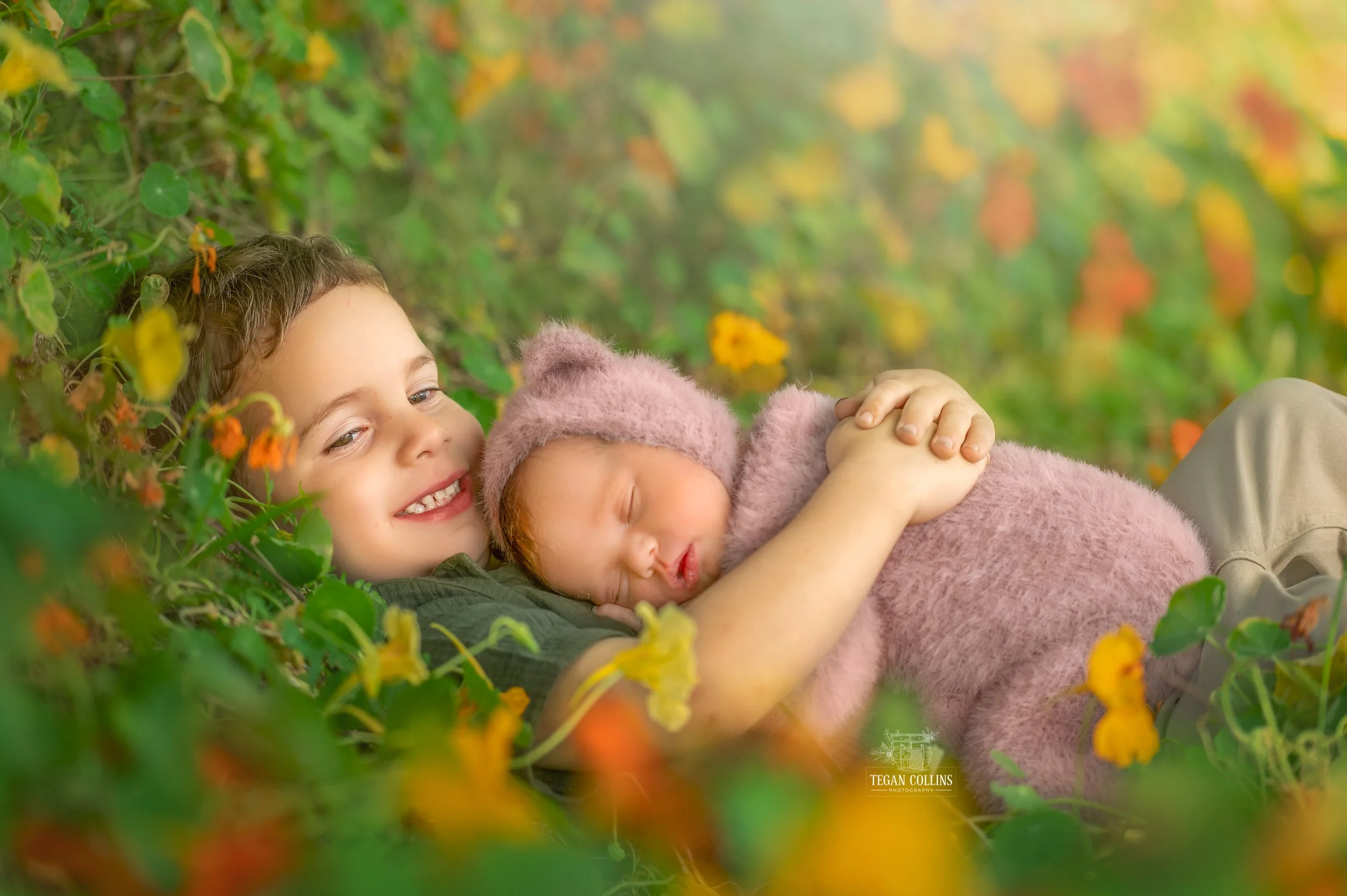 A big brother cuddles his newborn baby sister among the flowers. 