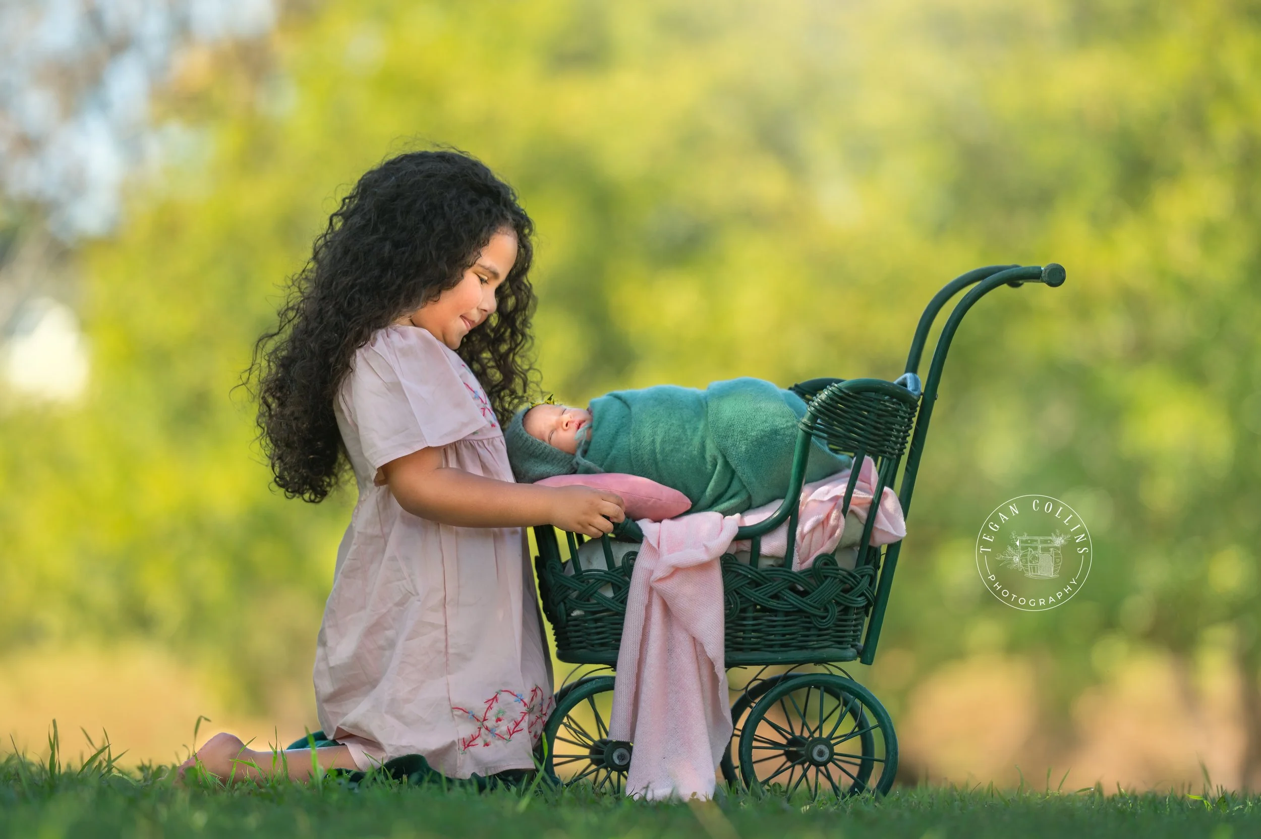A big sisters kneels beside a dolls pram while her newborn baby sister sleeps in the pram. The baby sister is wrapped in a green wrap with a pink blanket underneath her that matches her sisters pink dress.
