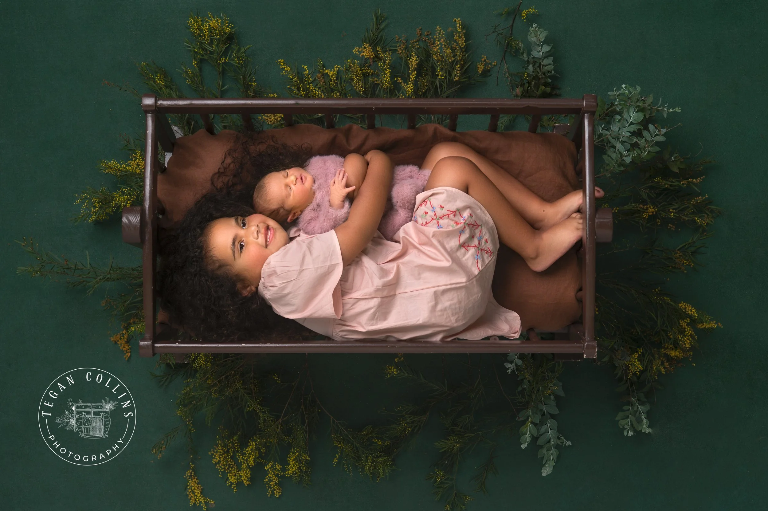 A green backdrop behind a big sister cuddling her newborn baby sister in a vintage cot. Both girls are wearing pink and wattle pokes out from under the cradle