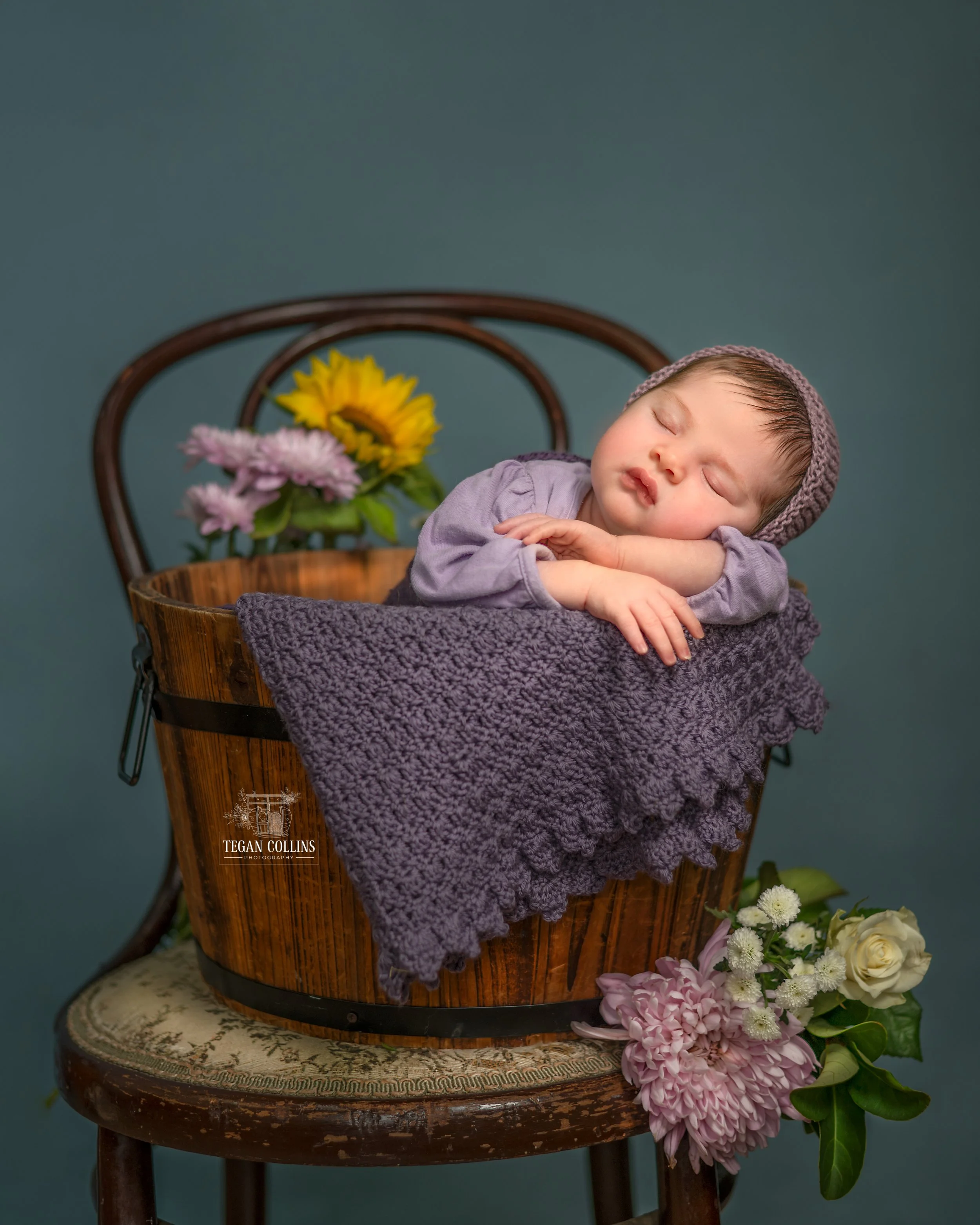 A newborn baby sleeps in an apple bucket amidst some spring flowers.