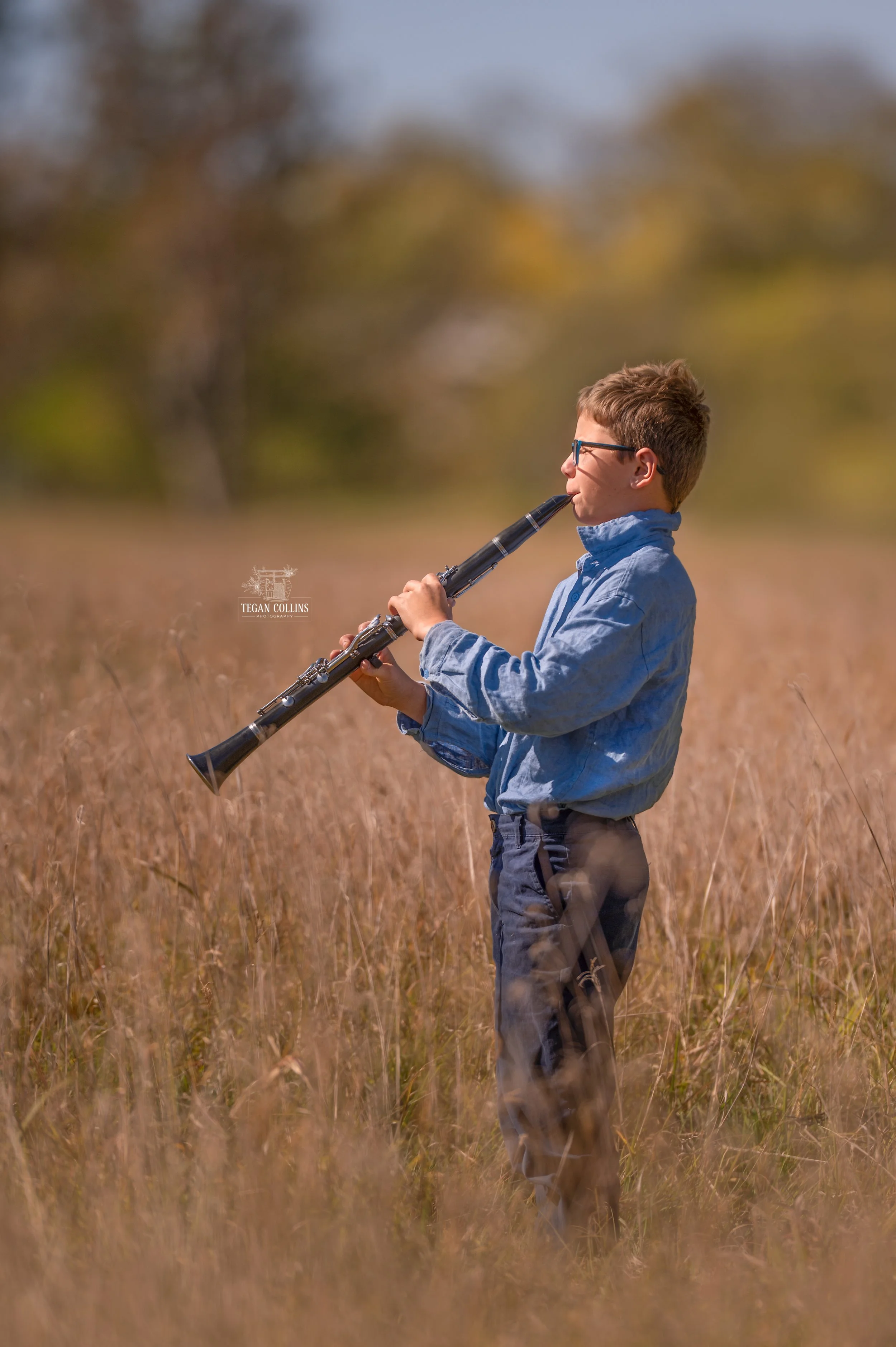 An Almost teen boy faces the sun in a field of winter grass and plays his clarinet. The boy is wearing a blue shirt and navy blue pants