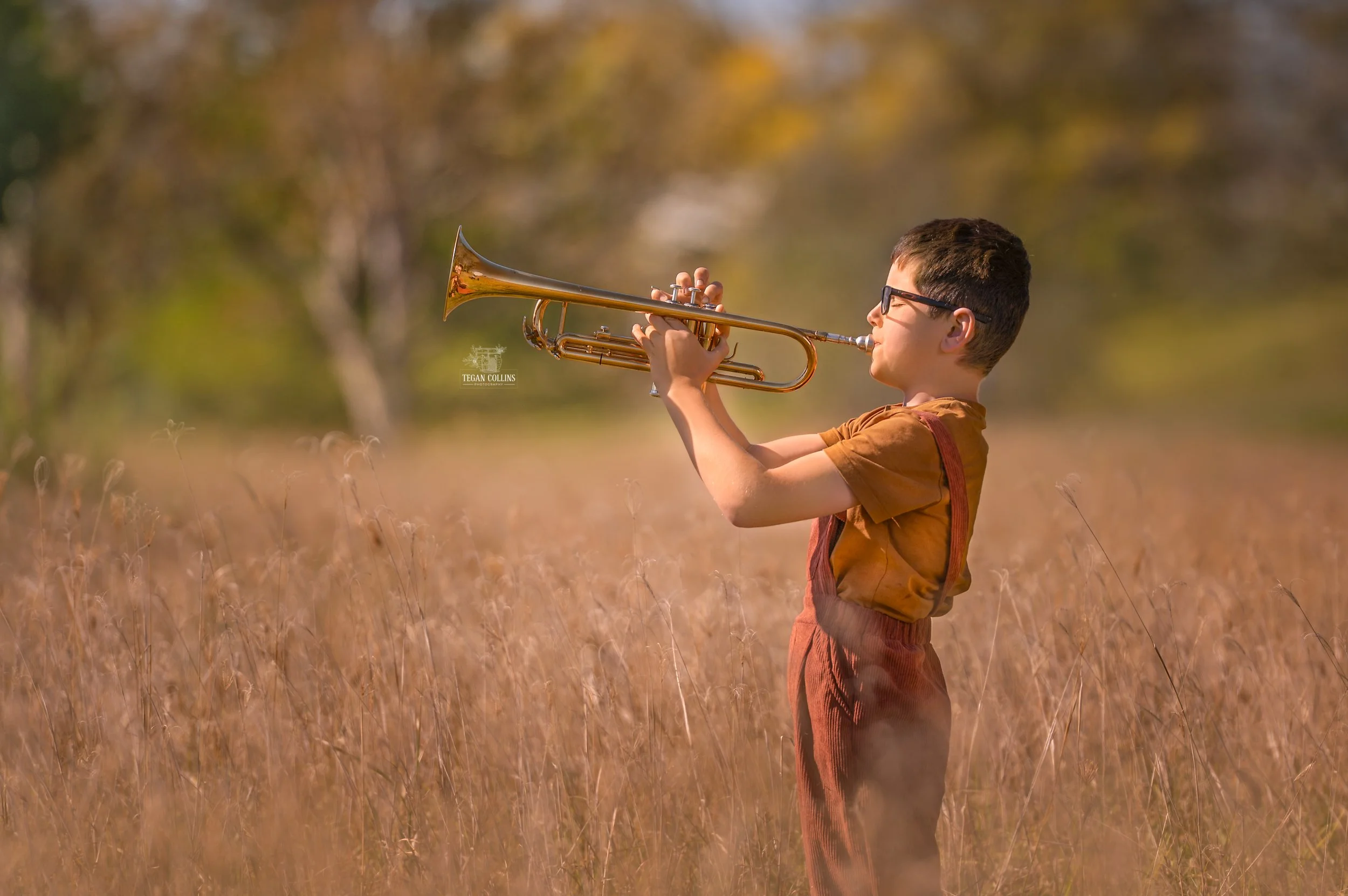 A young boy in a field of winter grass plays his trumpet. The boy is wearing a burnt orange t-shirt with brown overalls. The sun shines brightly on his face.