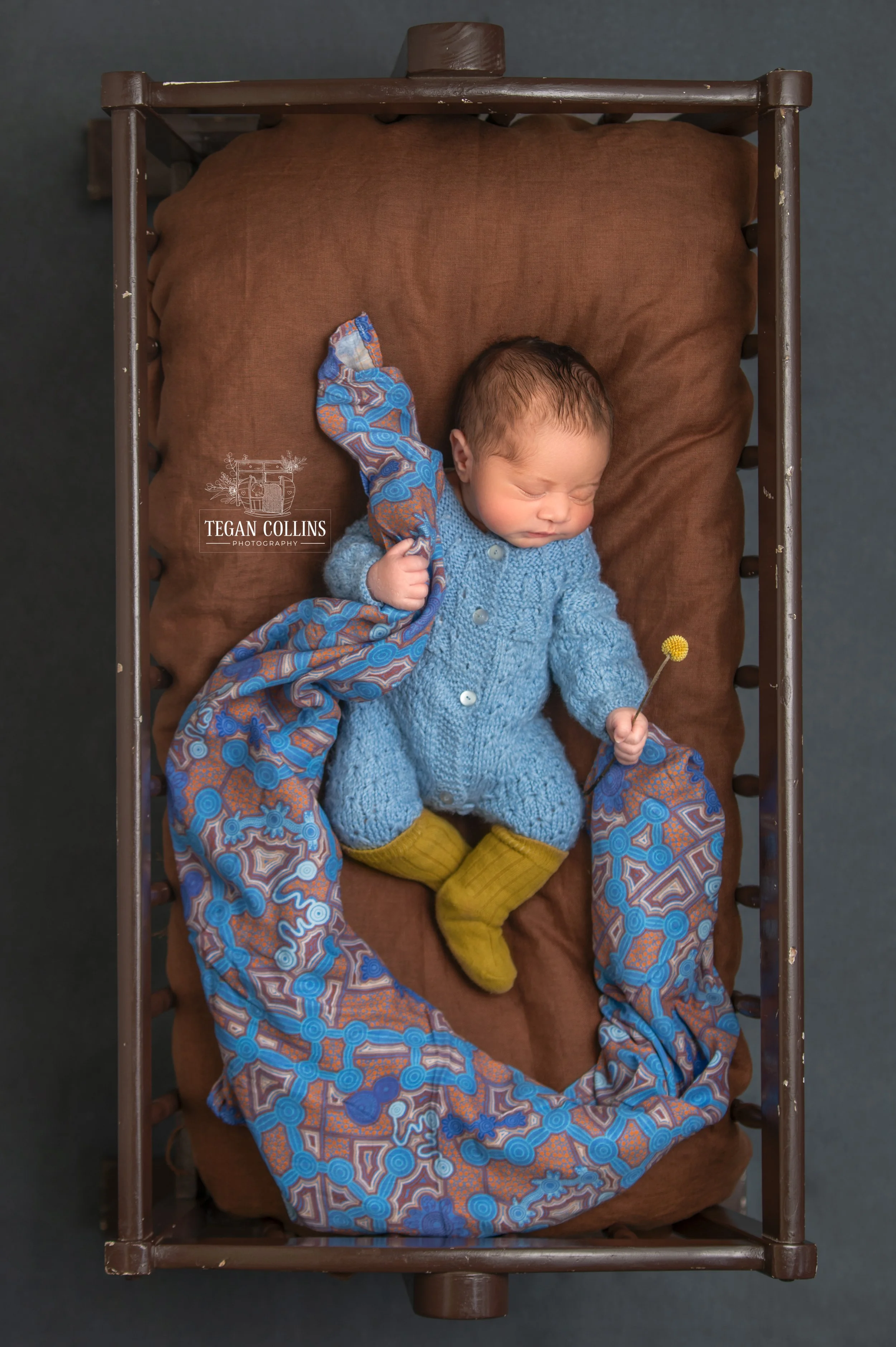 A crib where a newborn baby is seen sleeping and wrapping around him he is holding an aboriginal decorated blanket. 