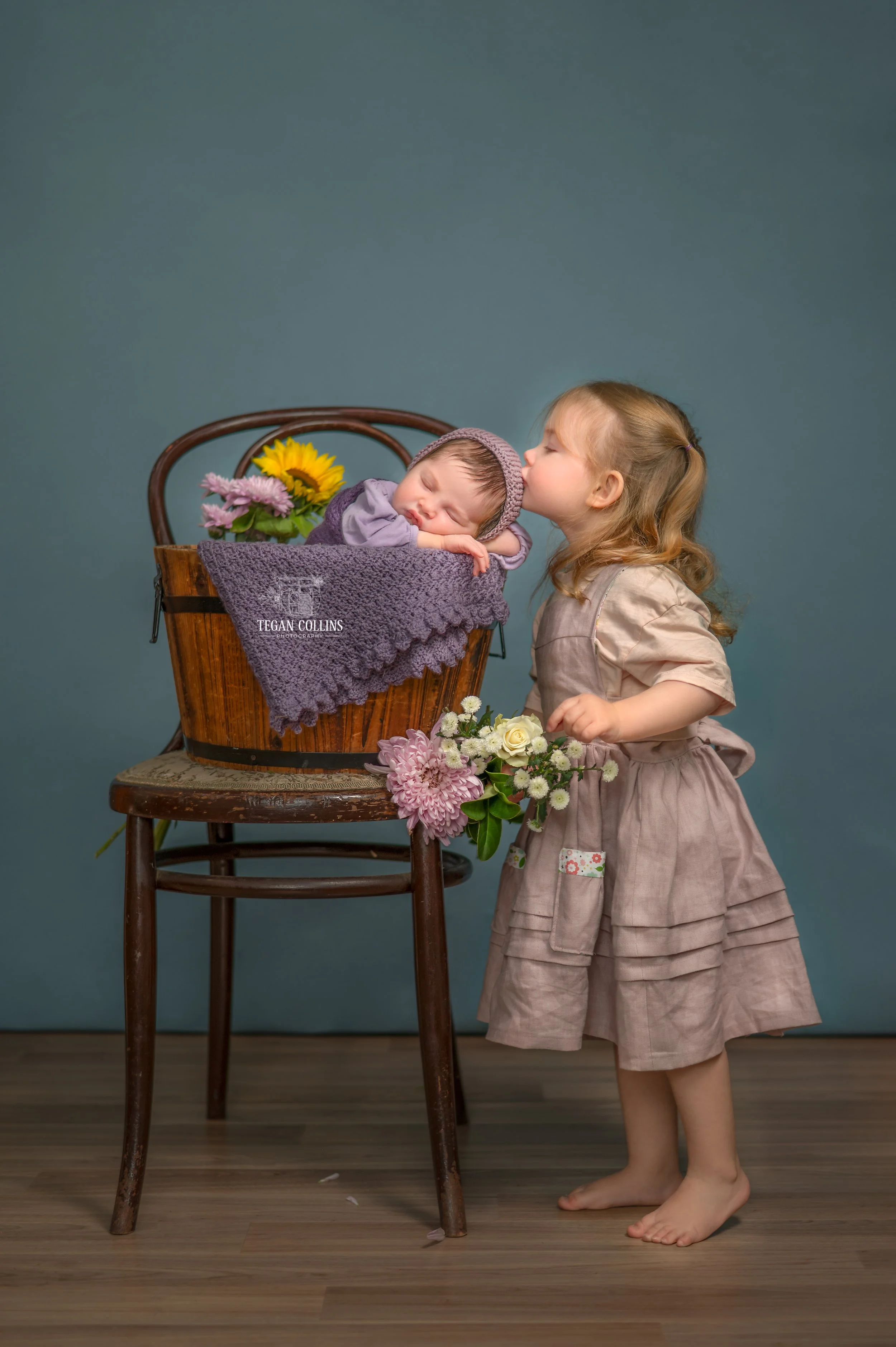 A big sister stretches up on her tippy toes to kiss her baby sisters head. 