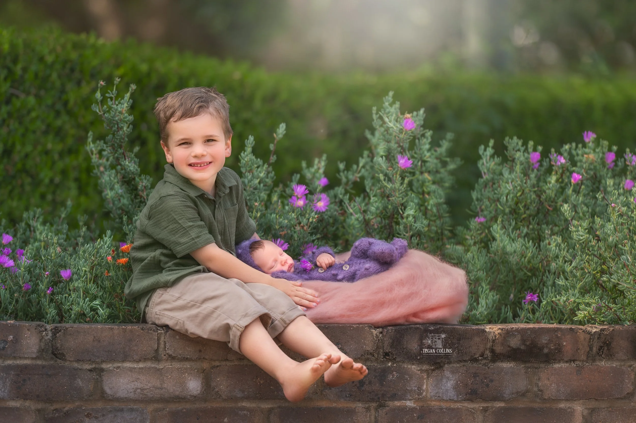 A little boy sits on a brick wall holding onto his new little sister who is wearing purple. 