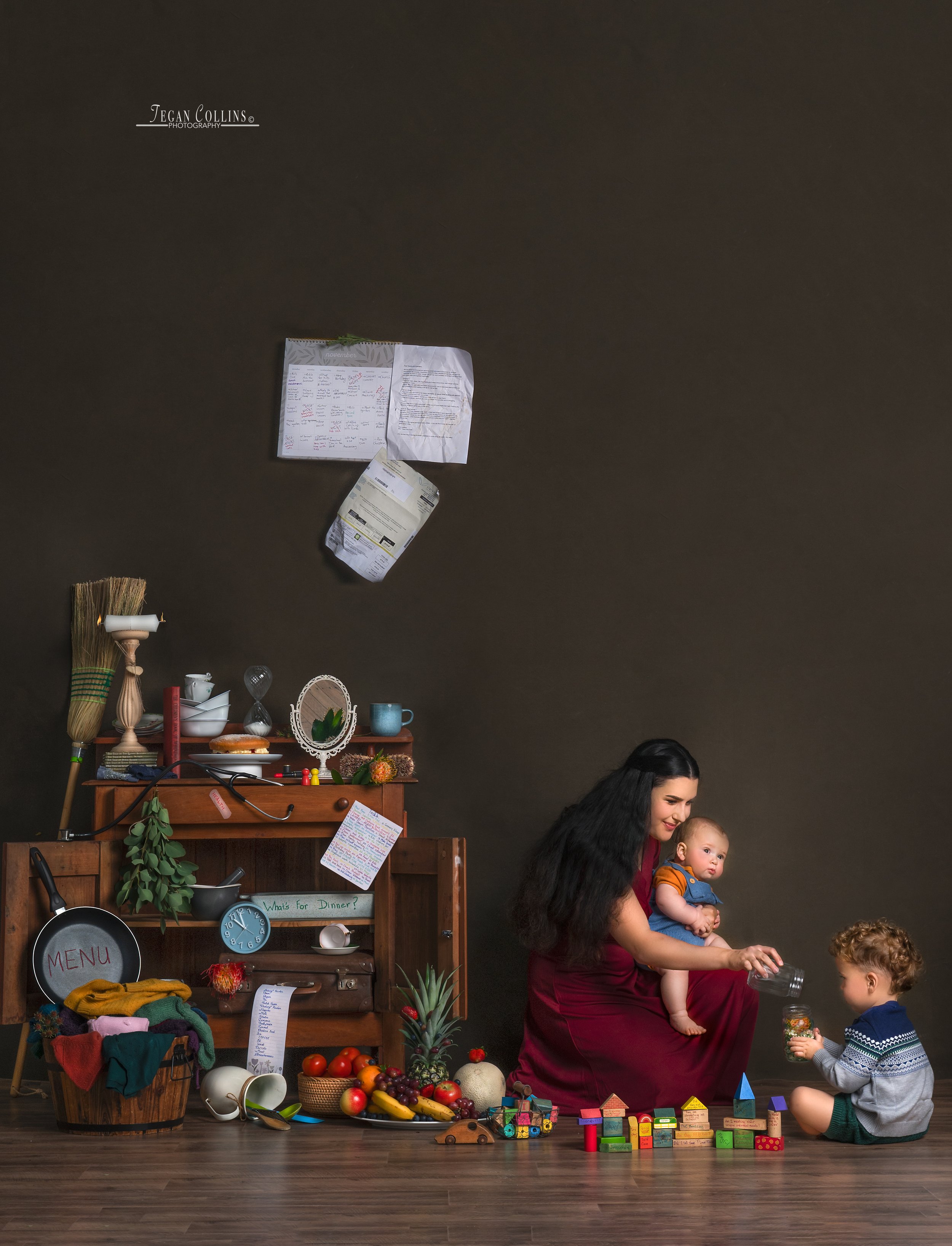 A woman sitting on the floor holding a baby with another young boy in front of her, in a room with a dark wall and a wooden shelf filled with various kitchen and decorative items, colorful fruits, and children's toys. Showing the weight of motherhood