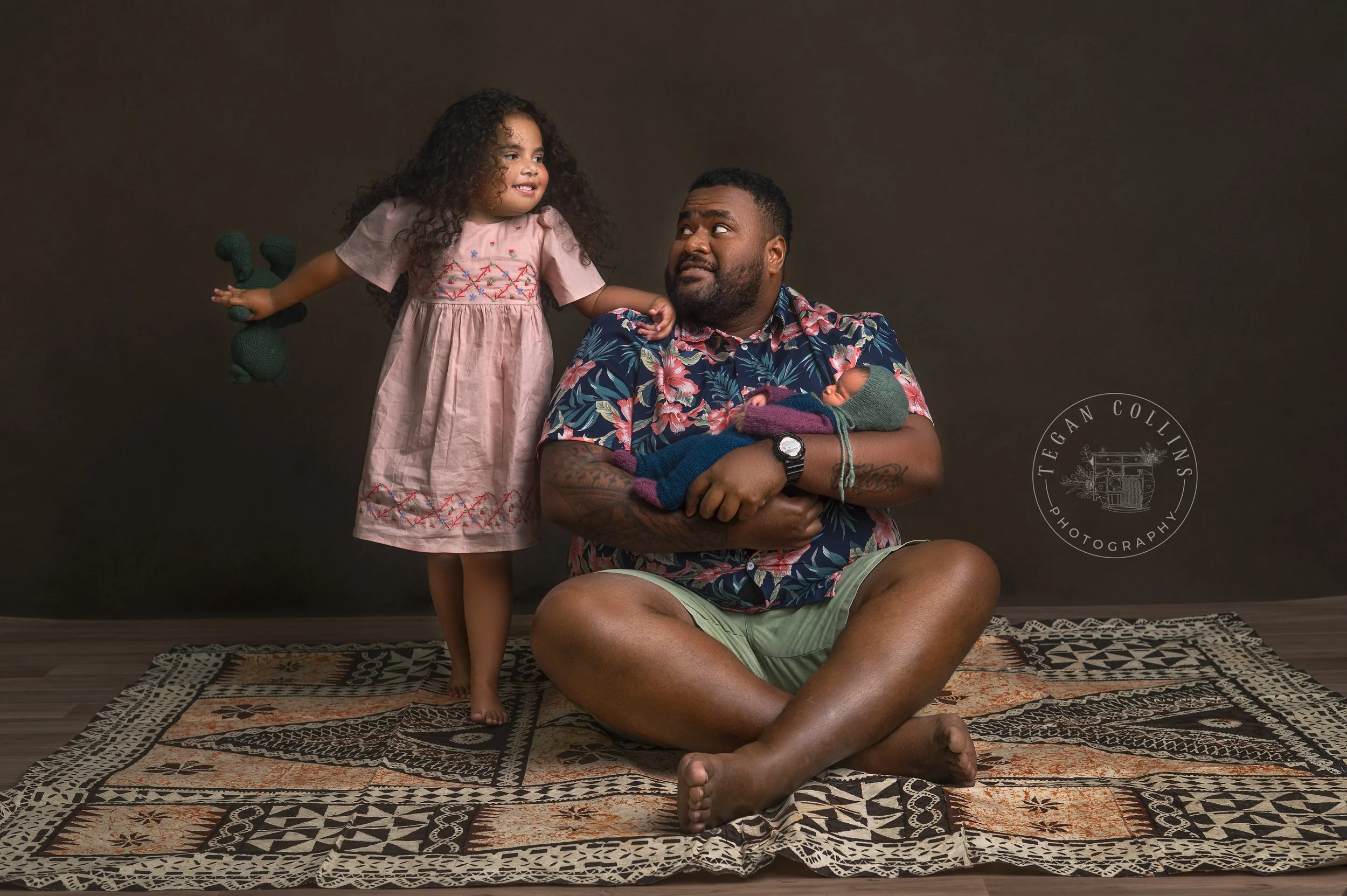 A dad is holding his newborn daughter on his traditional Fijian rug  while his big daughter stands beside him.