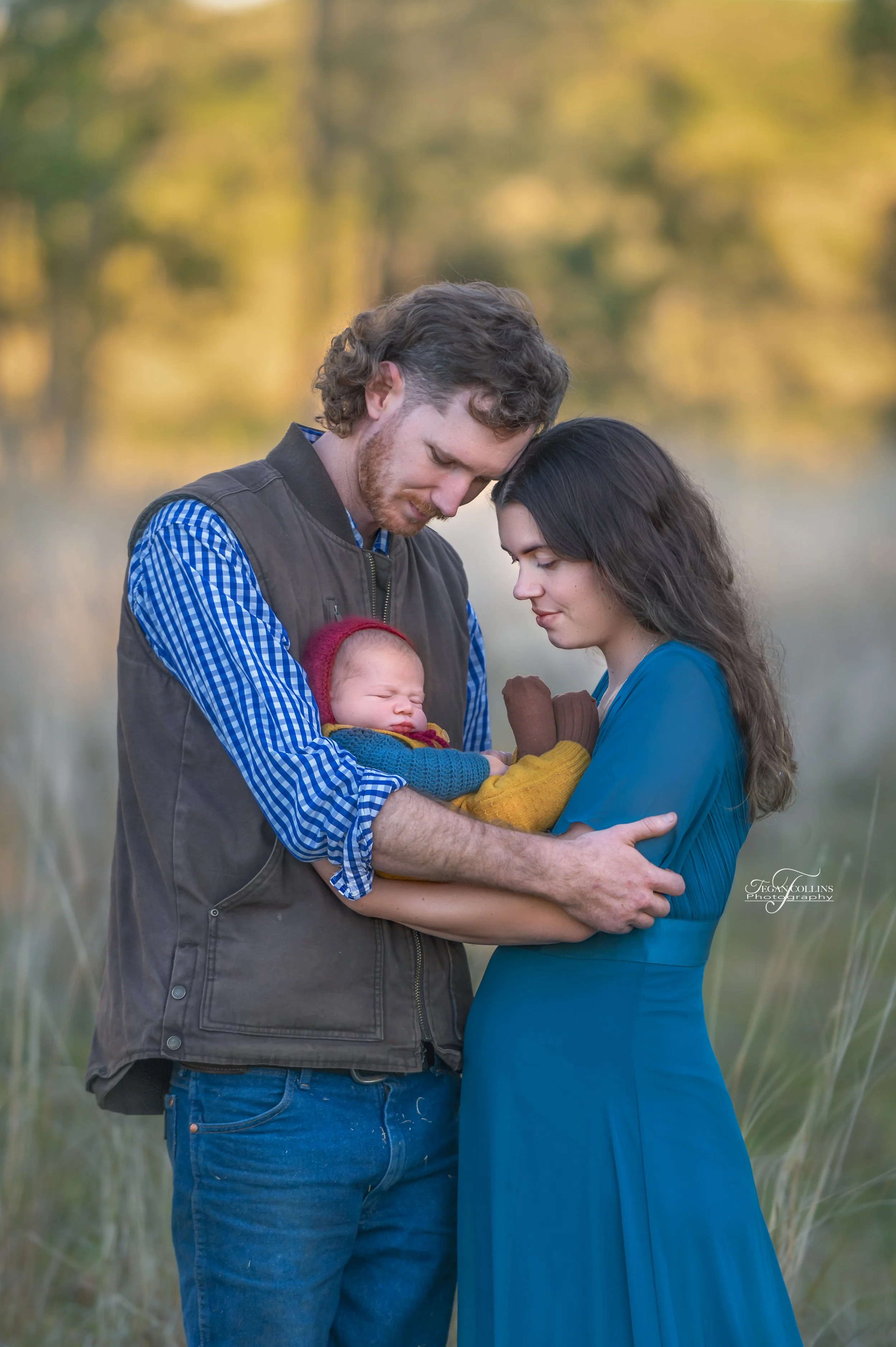 A mum and dad snuggle their newborn close. The background is dads family farm and property