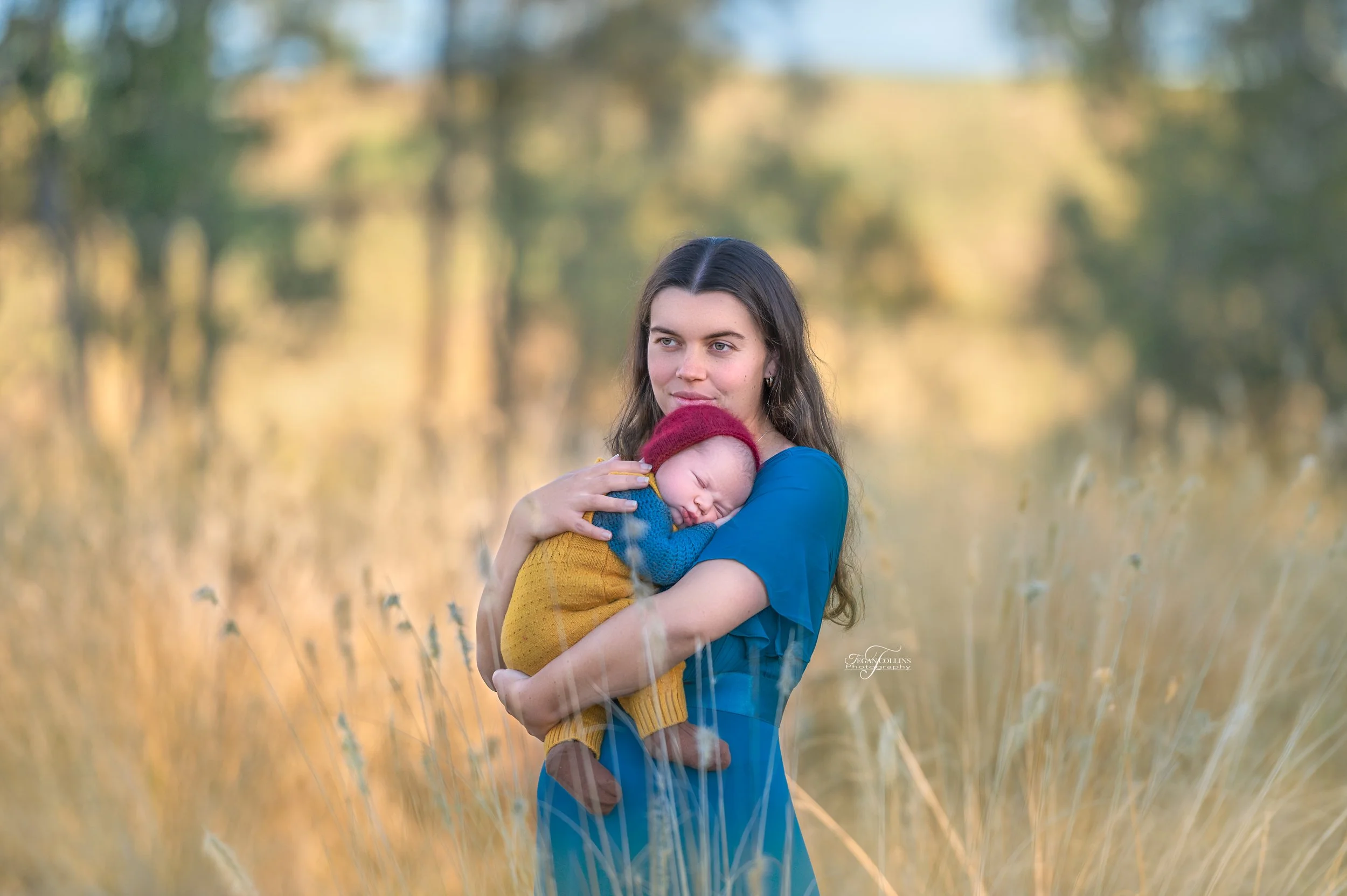 A mum wearing a striking blue dress snuggles her newborn baby who is wearing yellow overalls over a blue knitted suit and brown socks. on their family farm. 