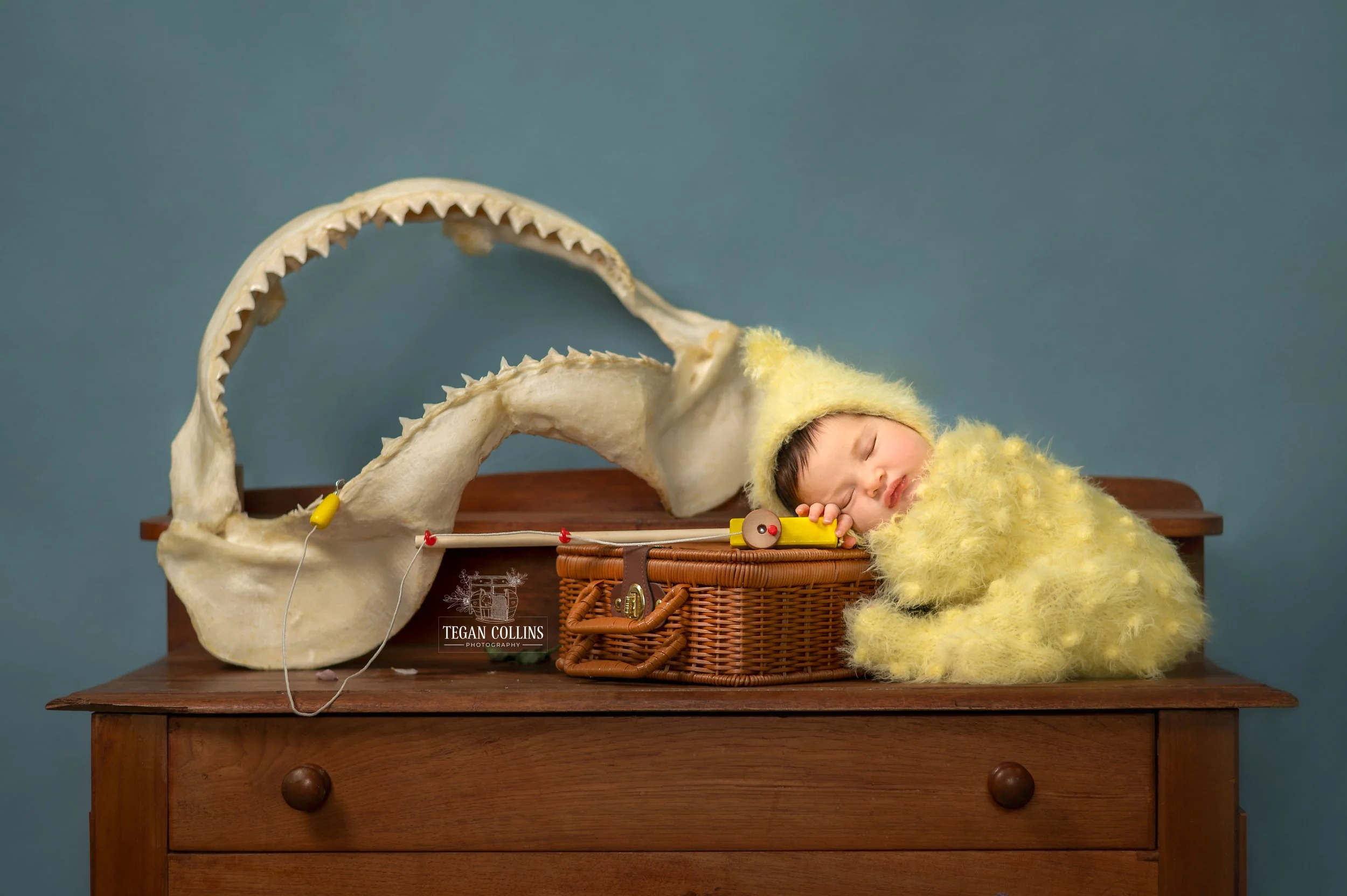 A baby girl wearing a yellow knitted bodysuit sits along an antique dresser holding a little fishing rod. behind her is a set of bull shark jaws.