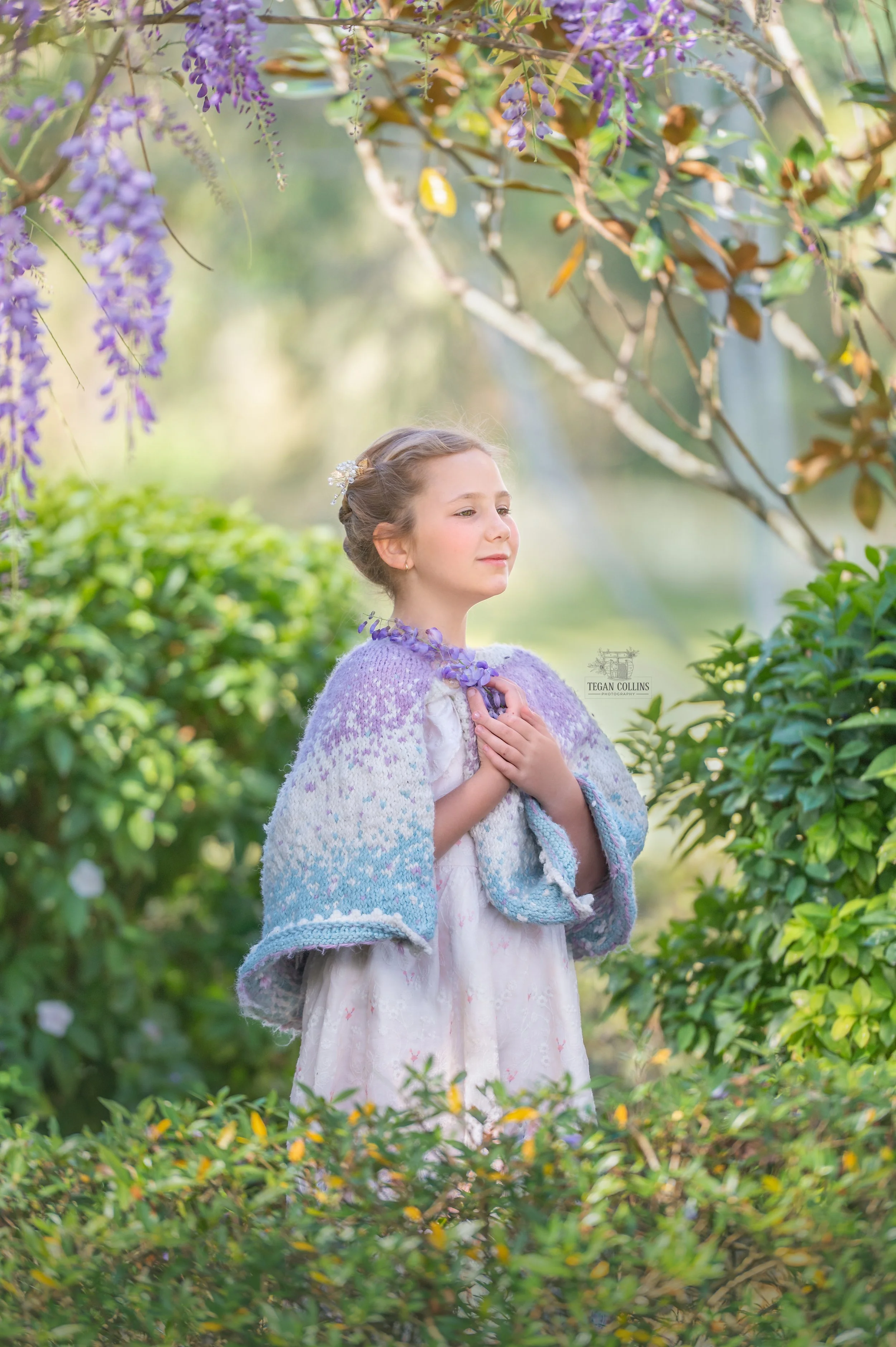 A young girl stands peacefully under purple wisteria. the girl is wearing a knitted caplet that matches the colour of the wisteria.
