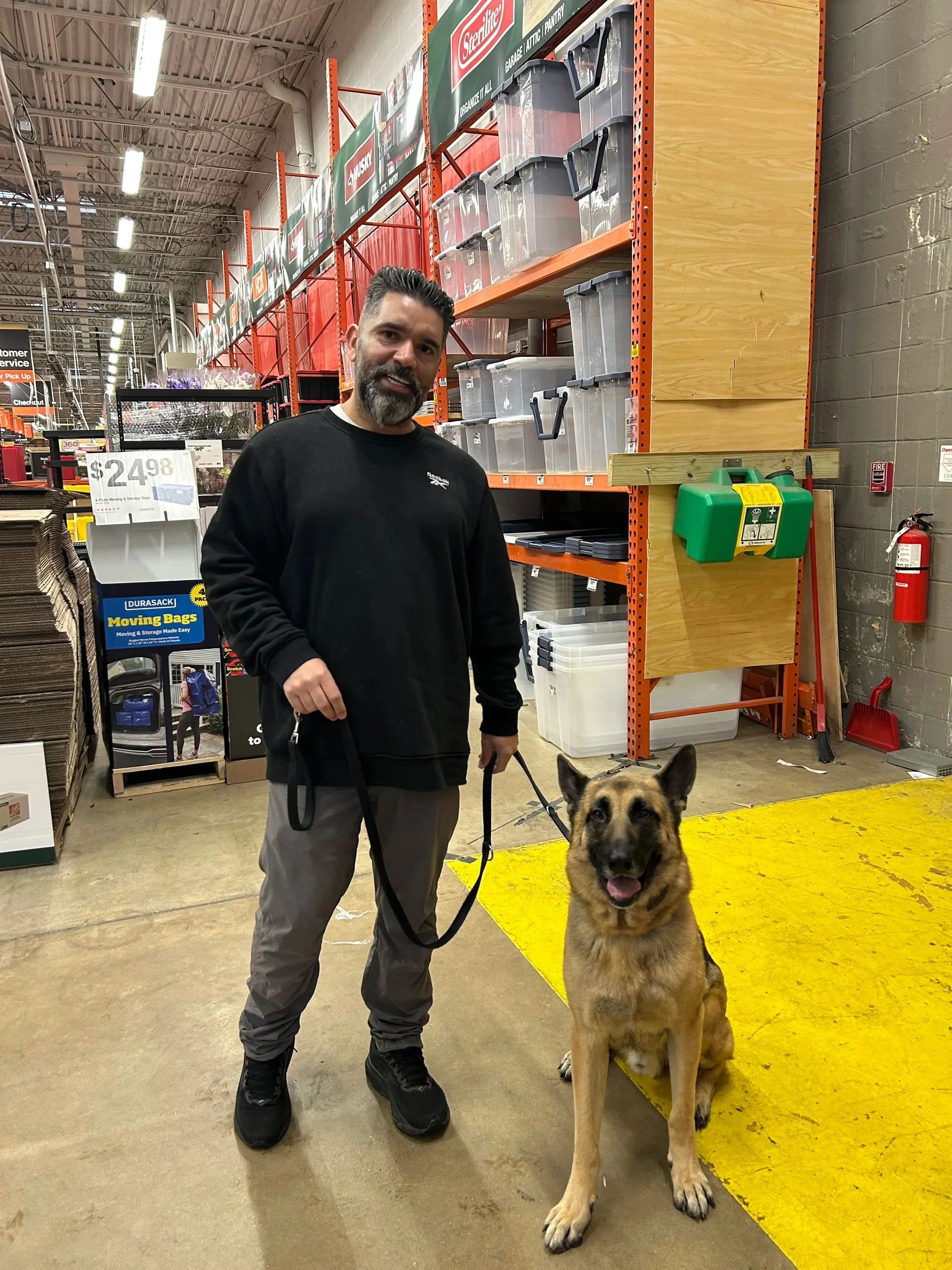 A man with dark hair and a beard, wearing black clothing, standing indoors next to a large German Shepherd dog on a leash. The man is smiling and the dog has its tongue out, sitting on a yellow mat. The background includes metal shelving with storage bins, signs, and a fire alarm on a concrete wall.