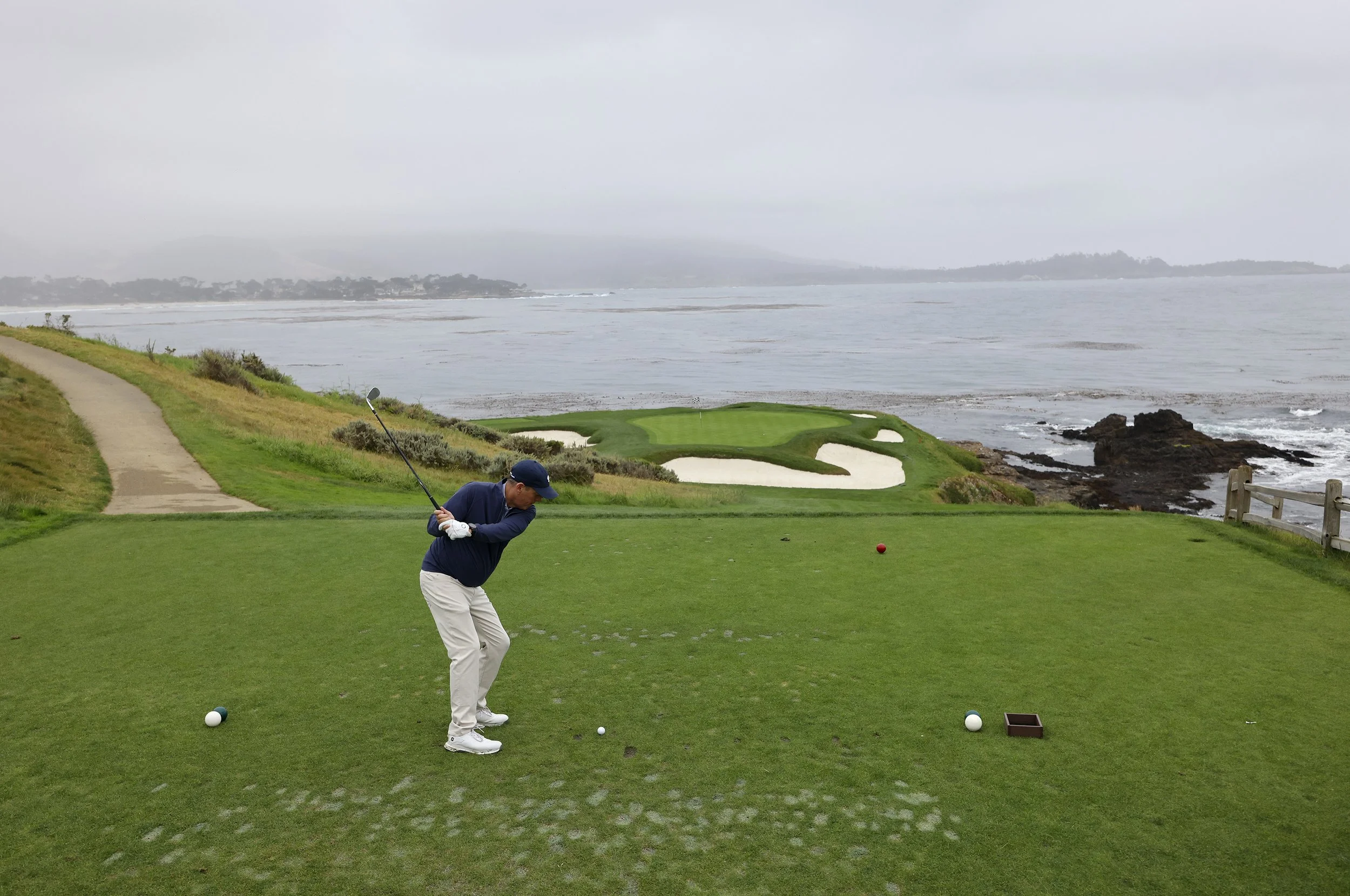 A golfer in a dark blue shirt and white pants tees off on the famouse 7th hole at Pebble Beach Golf Links.