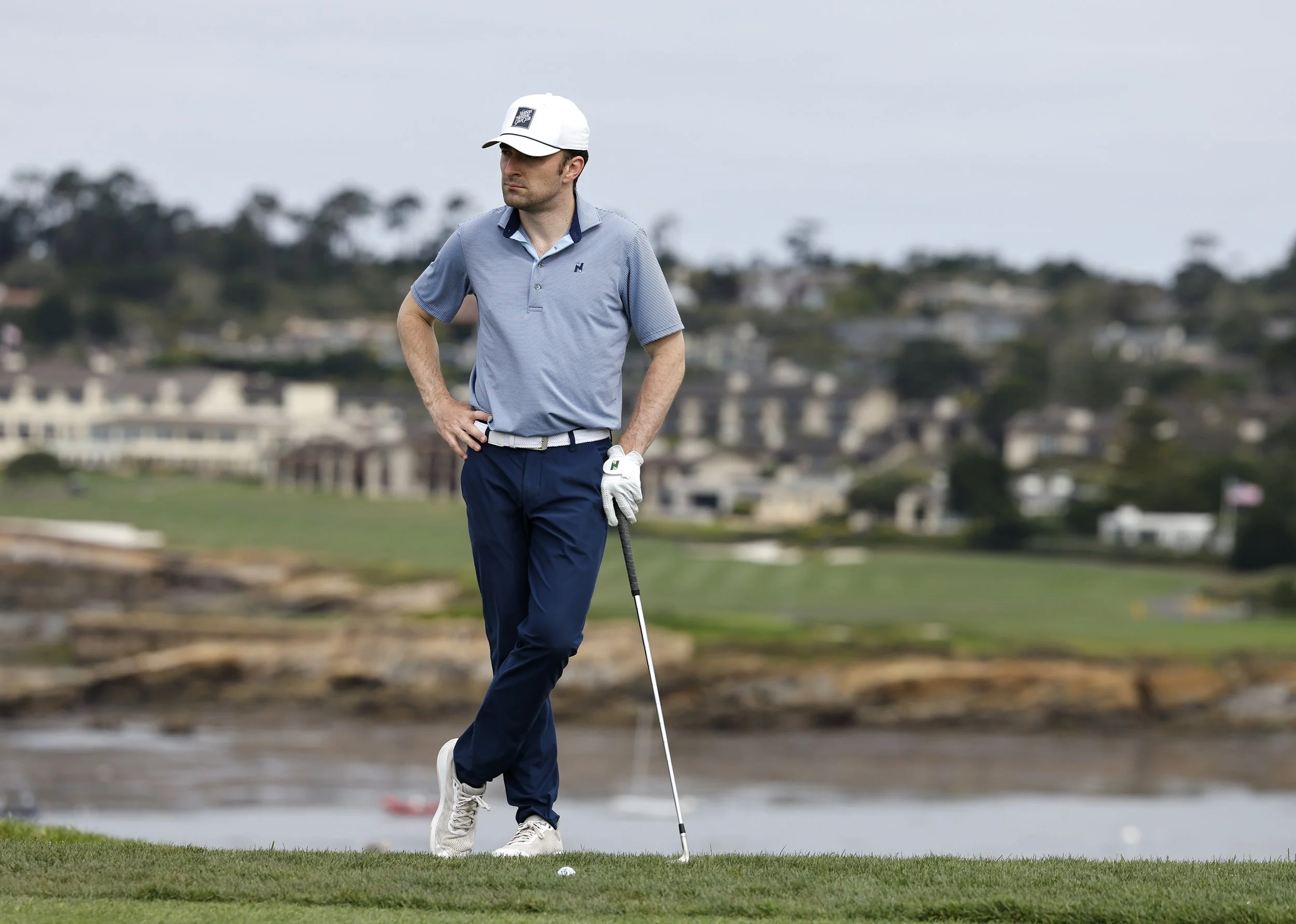 A man in a blue striped golf shirt, blue pants, and a white cap is standing on a golf course. He is holding a golf club and appears to be preparing to take a shot, with a golf ball on the ground near him. The background features a blurred view of hou
