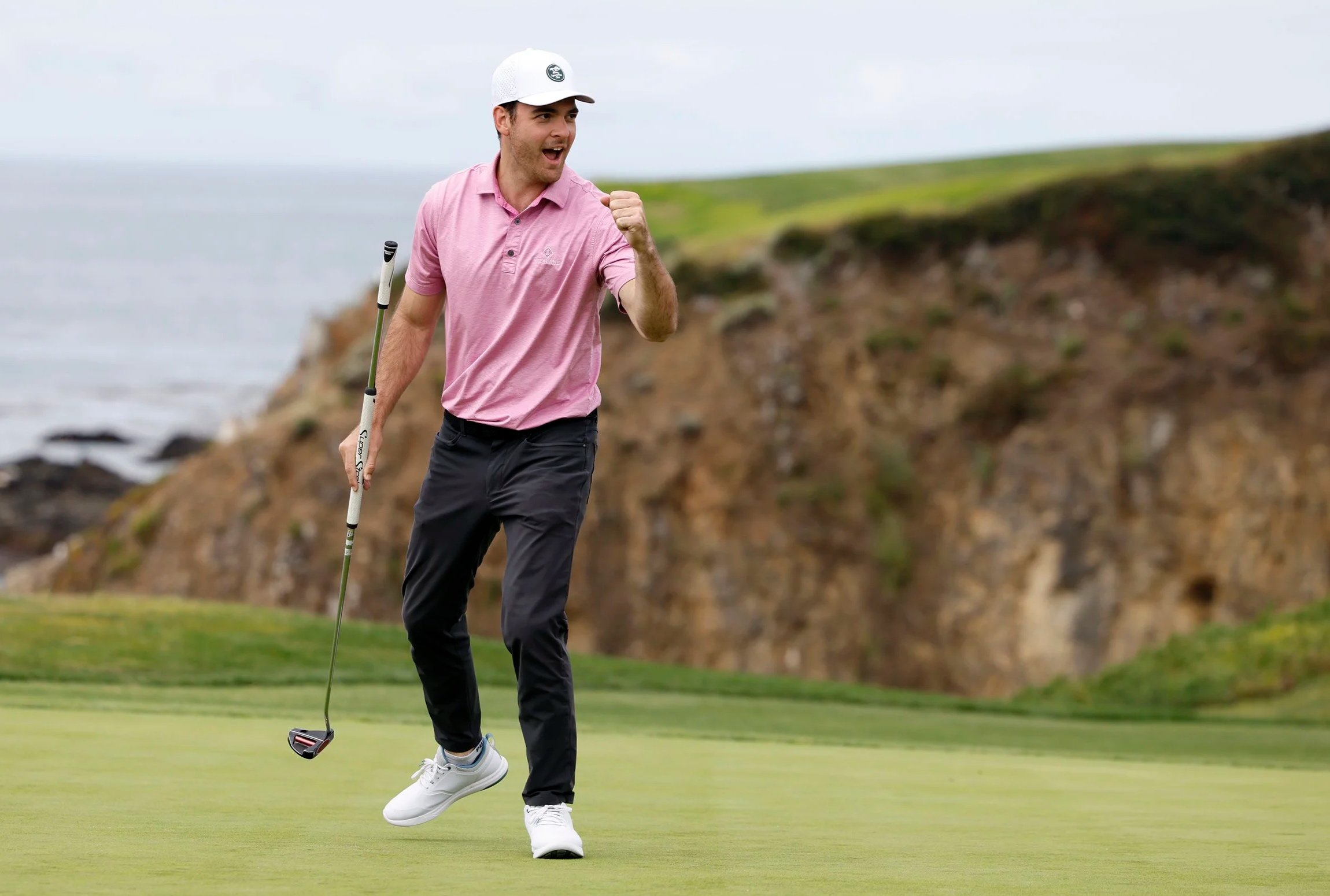 A man in a pink polo shirt, dark pants, and white shoes celebrating on a golf course, holding a golf club. He is smiling and raising his fist in victory with a seaside cliff in the background.