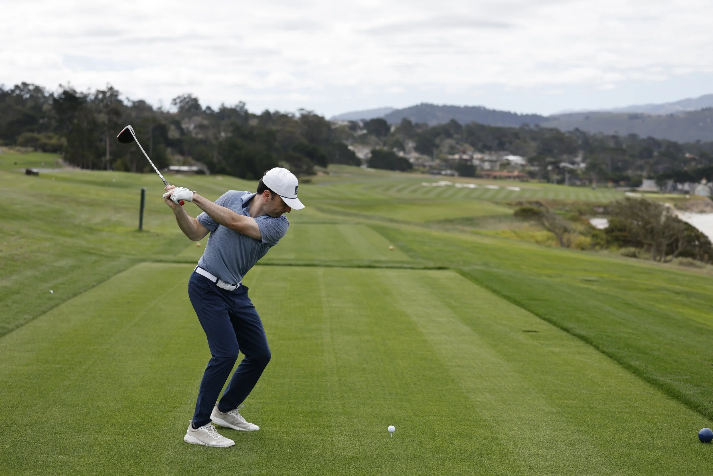 A man in a white cap and blue golf shirt swinging a golf club on a green golf course with rolling hills and trees in the background.