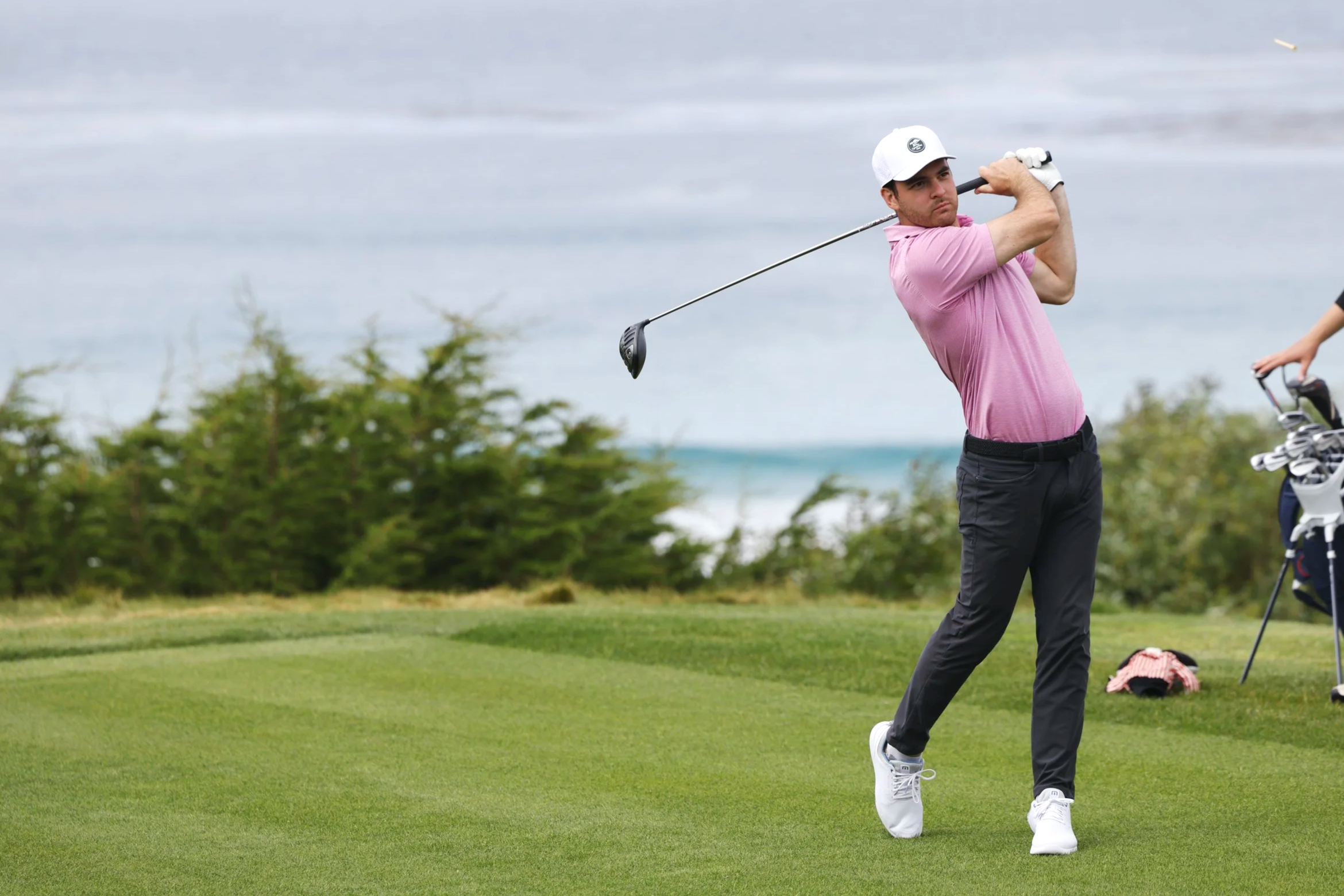 A man in a pink shirt, black pants, and white shoes swinging a golf club on a golf course with greenery and water in the background.