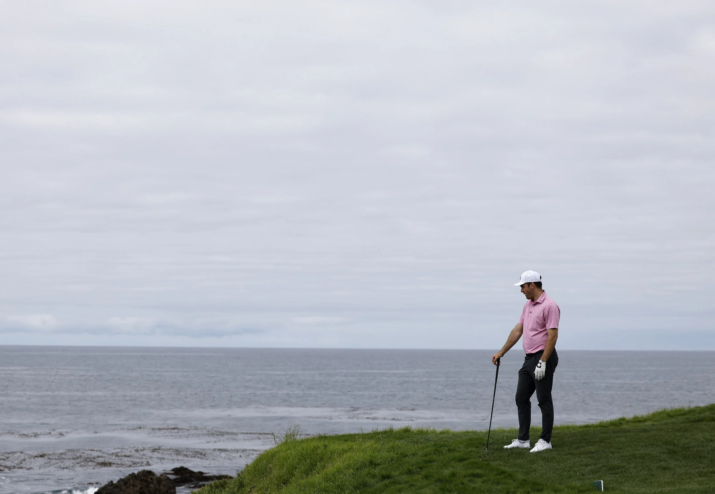 A man in a pink shirt, gray pants, and white shoes standing on a grassy area near the water, holding a golf club, looking down, with a cloudy sky background.