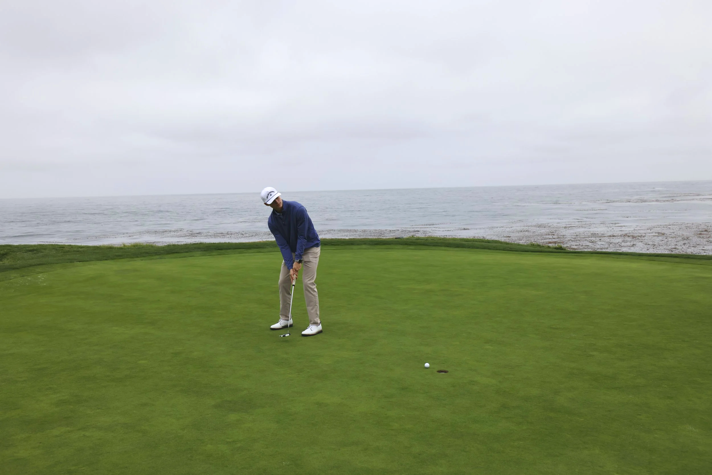 A man in a blue jacket and beige pants playing golf on a green course near a lake with cloudy sky.