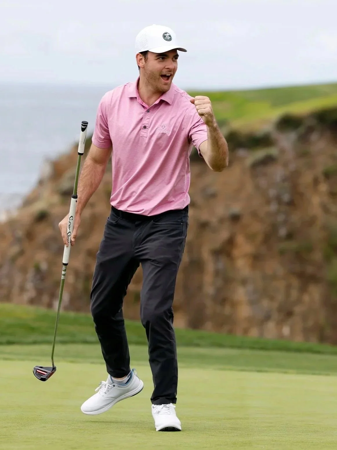 A man celebrating on a golf course, holding a golf club, wearing a pink polo shirt, black pants, white shoes, and a white cap, with a scenic landscape in the background.