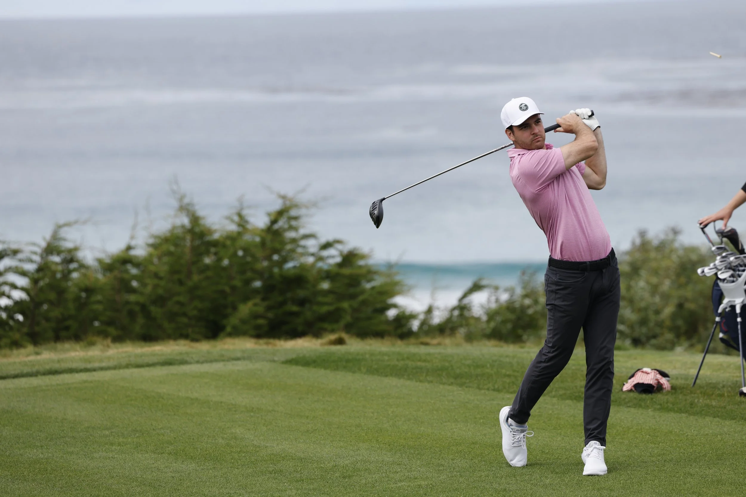 A man wearing a white cap, pink polo, and black pants is swinging a golf club on a golf course with greenery and water in the background on the 10th hole at Pebble Beach Golf Links